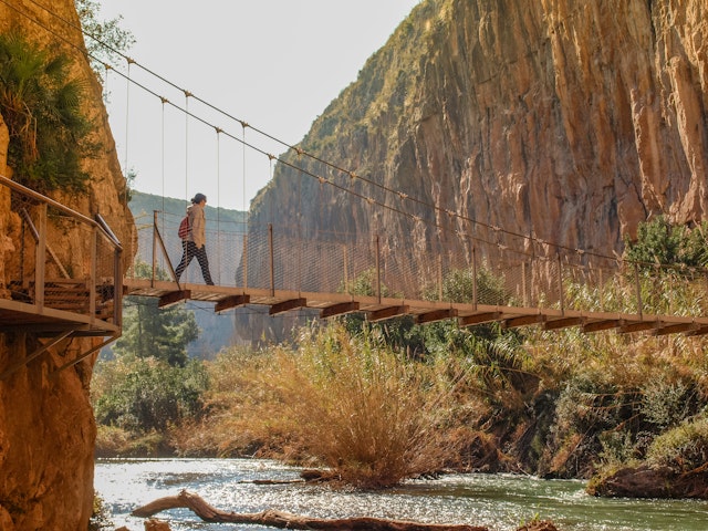 A hiker walks over a suspension bridge above a river, linking up two sides of a gorge