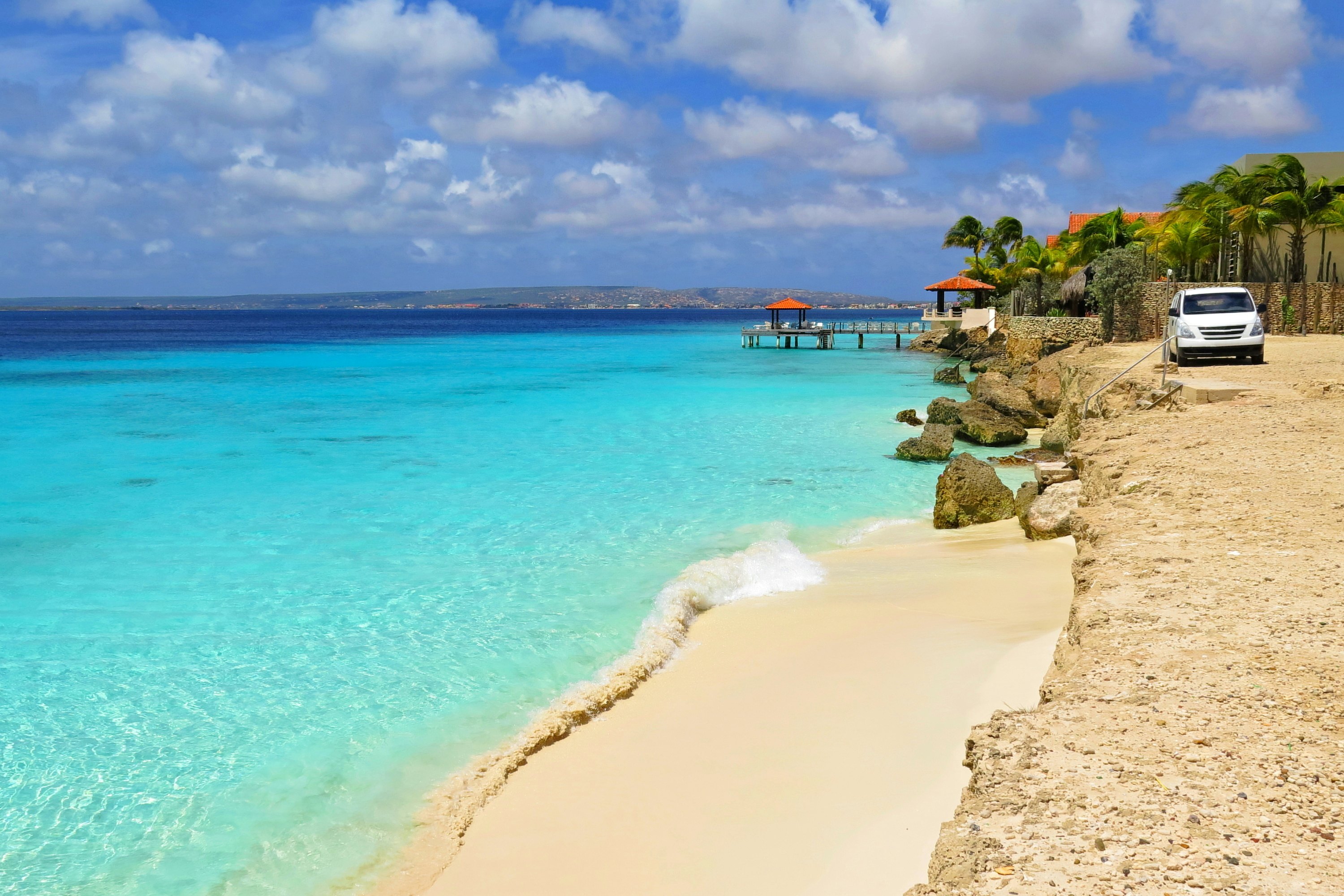 A car is parked on a bluff above a narrow sandy beach with shallow waters looking azure in the tropical sun, Aruba