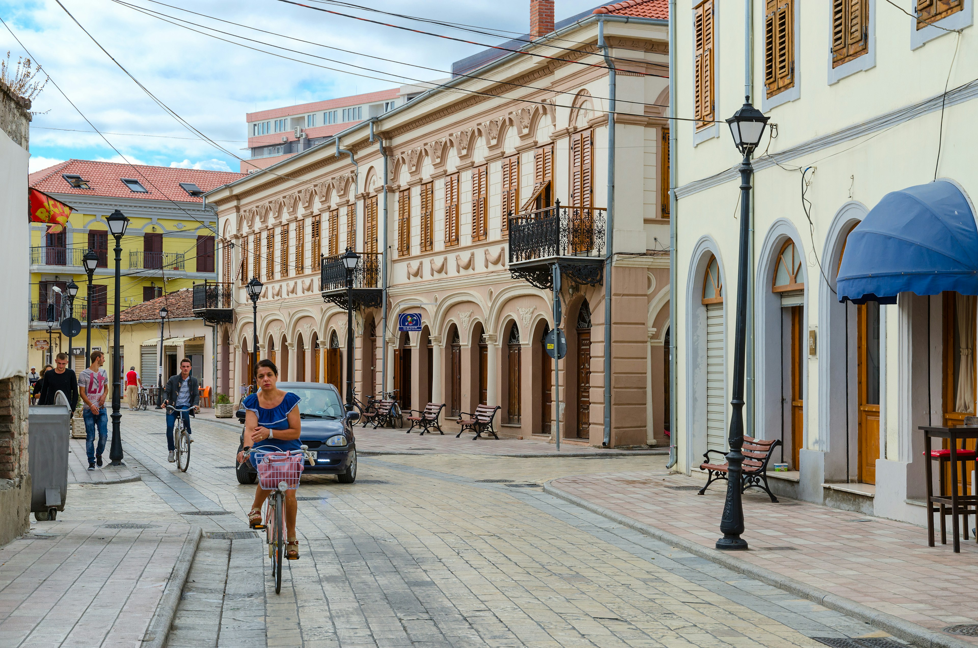 People ride bicycles and walk past elegant, historic buildings along street (Rruga G'juhadol) in center of Shkoder, Albania