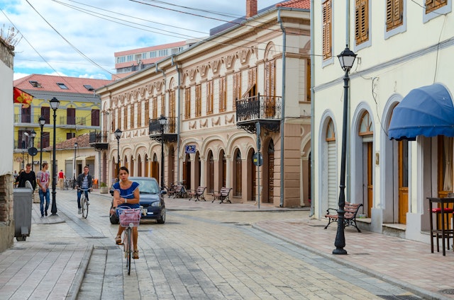 People ride bicycles and walk past elegant, historic buildings along street (Rruga G'juhadol) in center of Shkoder, Albania