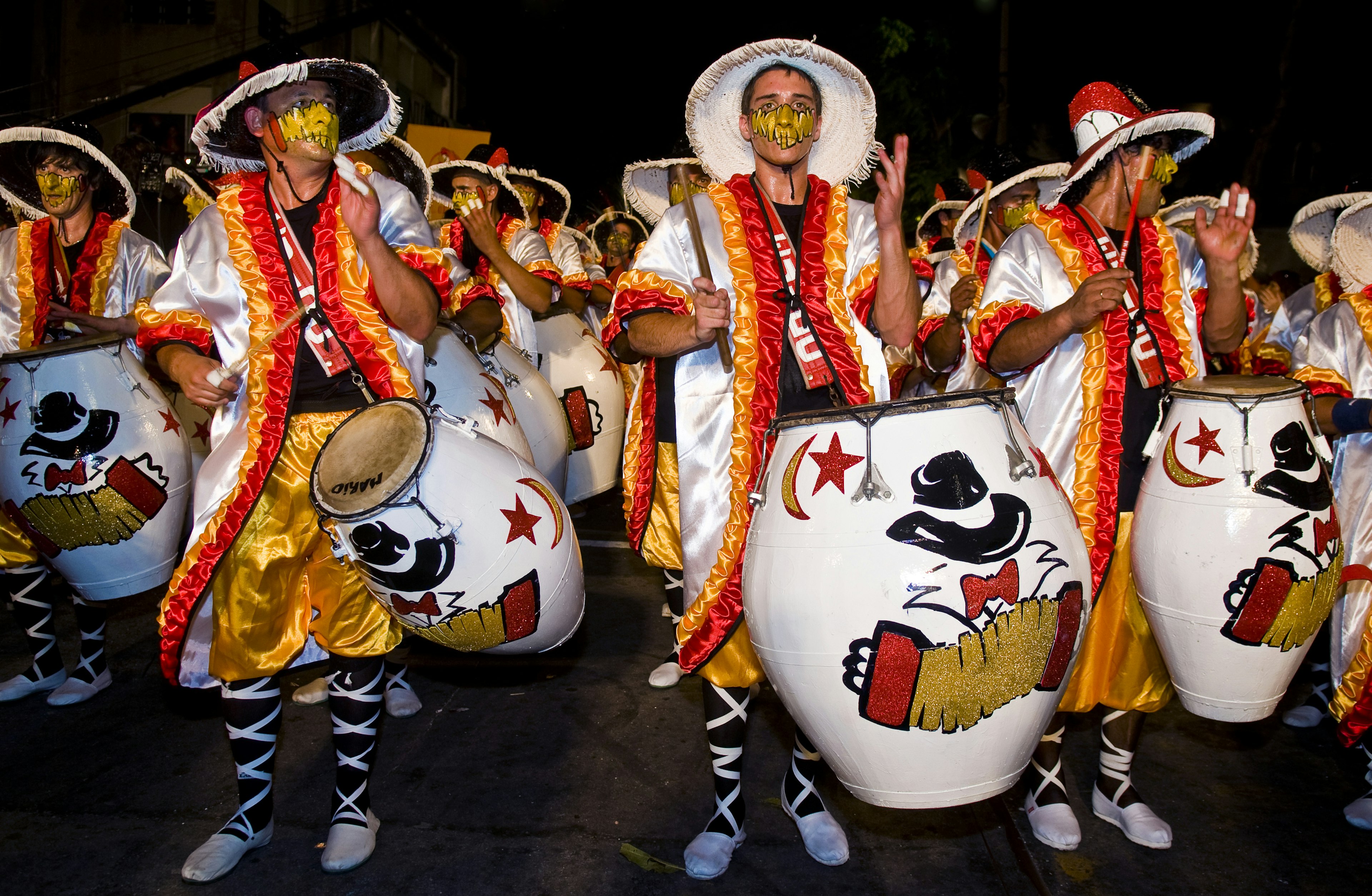 A group of people clad in orange, black and white robes play large white drums