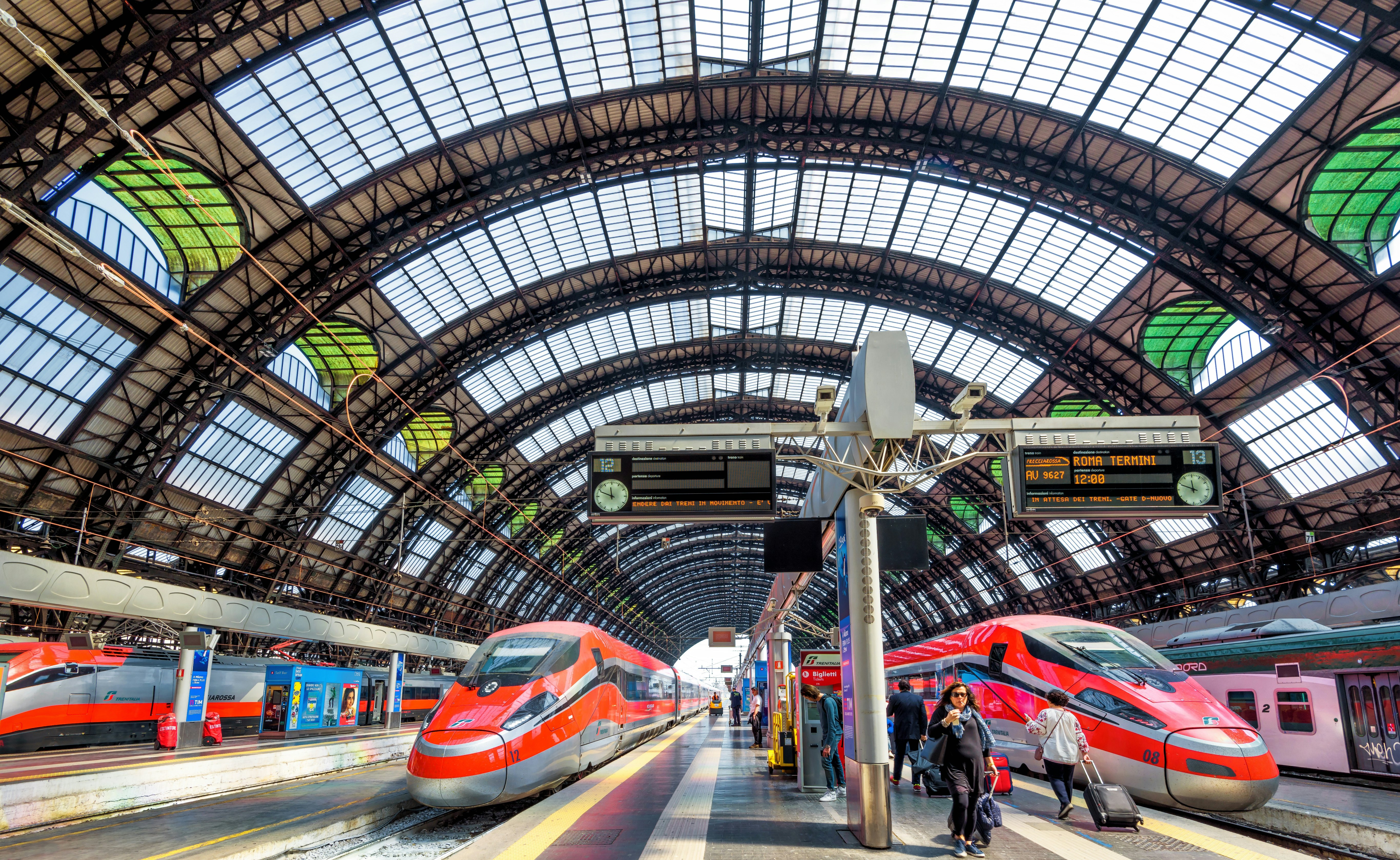 Two red high-speed trains wait at platfomrs in a grand station with an arched glass roof