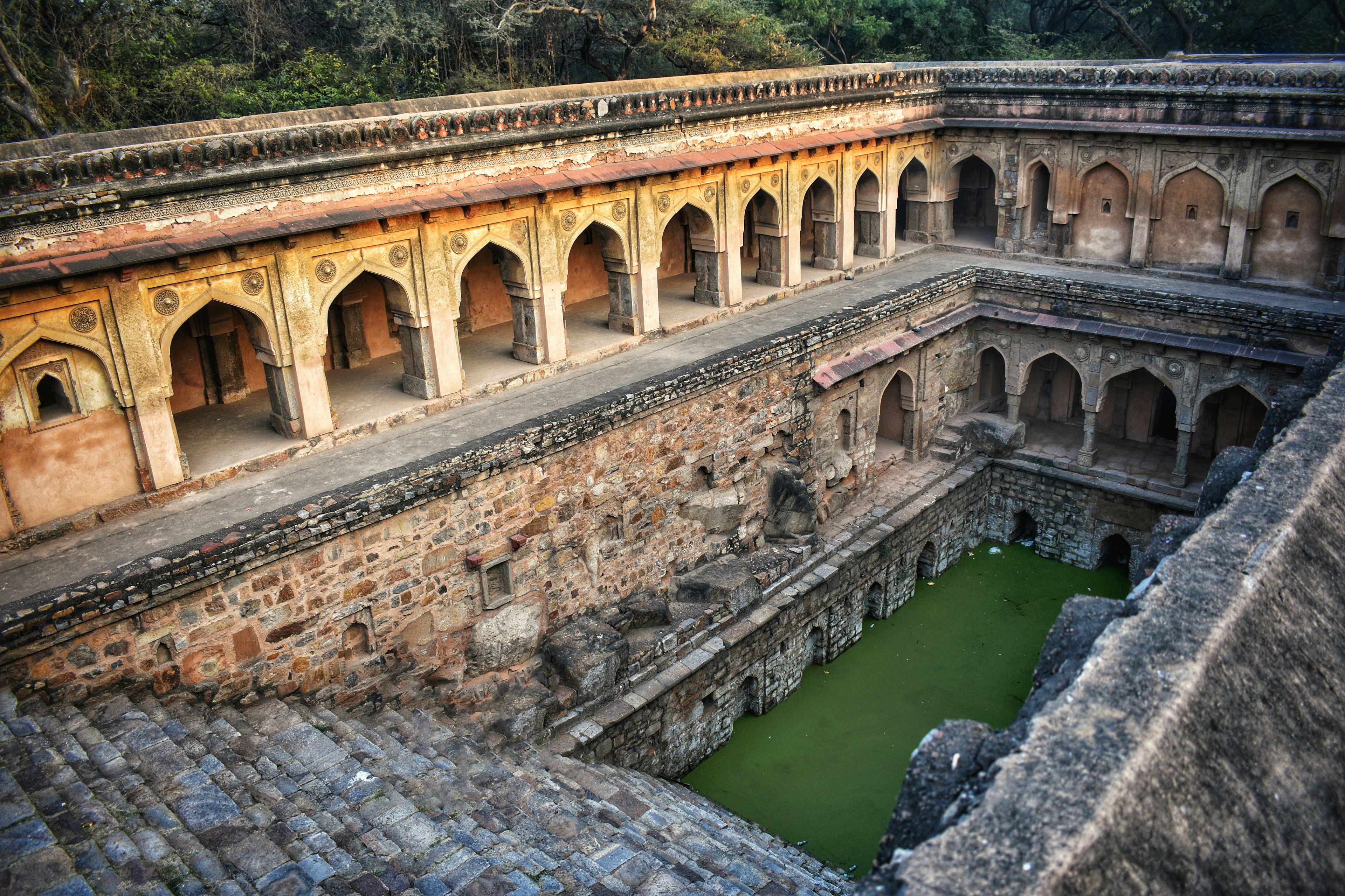 Stone stairs descend into the shallow waters of Rajon Ki Baoli stepwell in Delhi. The surrounding stone walls are tiered, with built-in archways and decorated with Islamic stucco motifs.