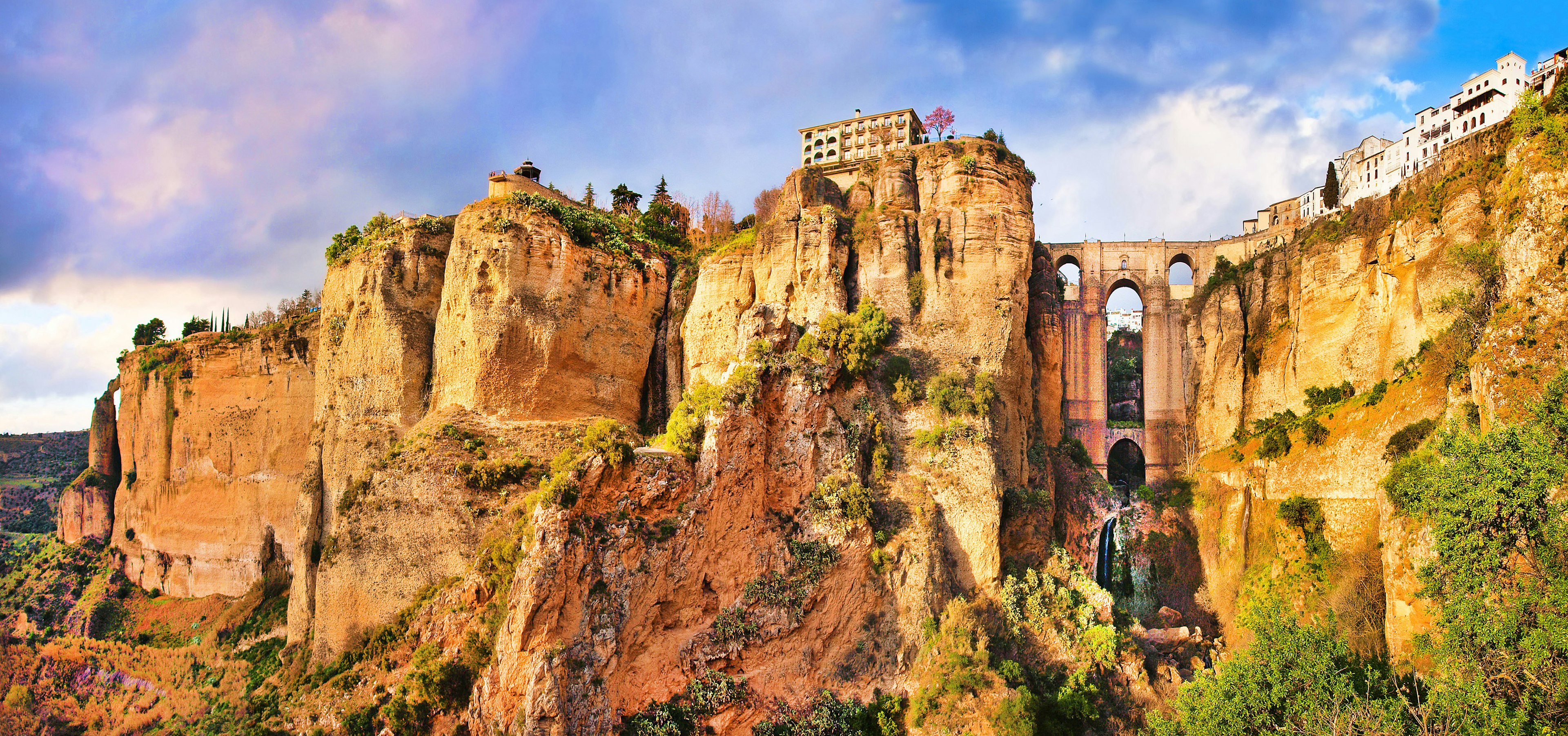 Looking up at an arched bridge dramatically suspended across a gorge between tall sand-coloured cliffs with houses on either side.