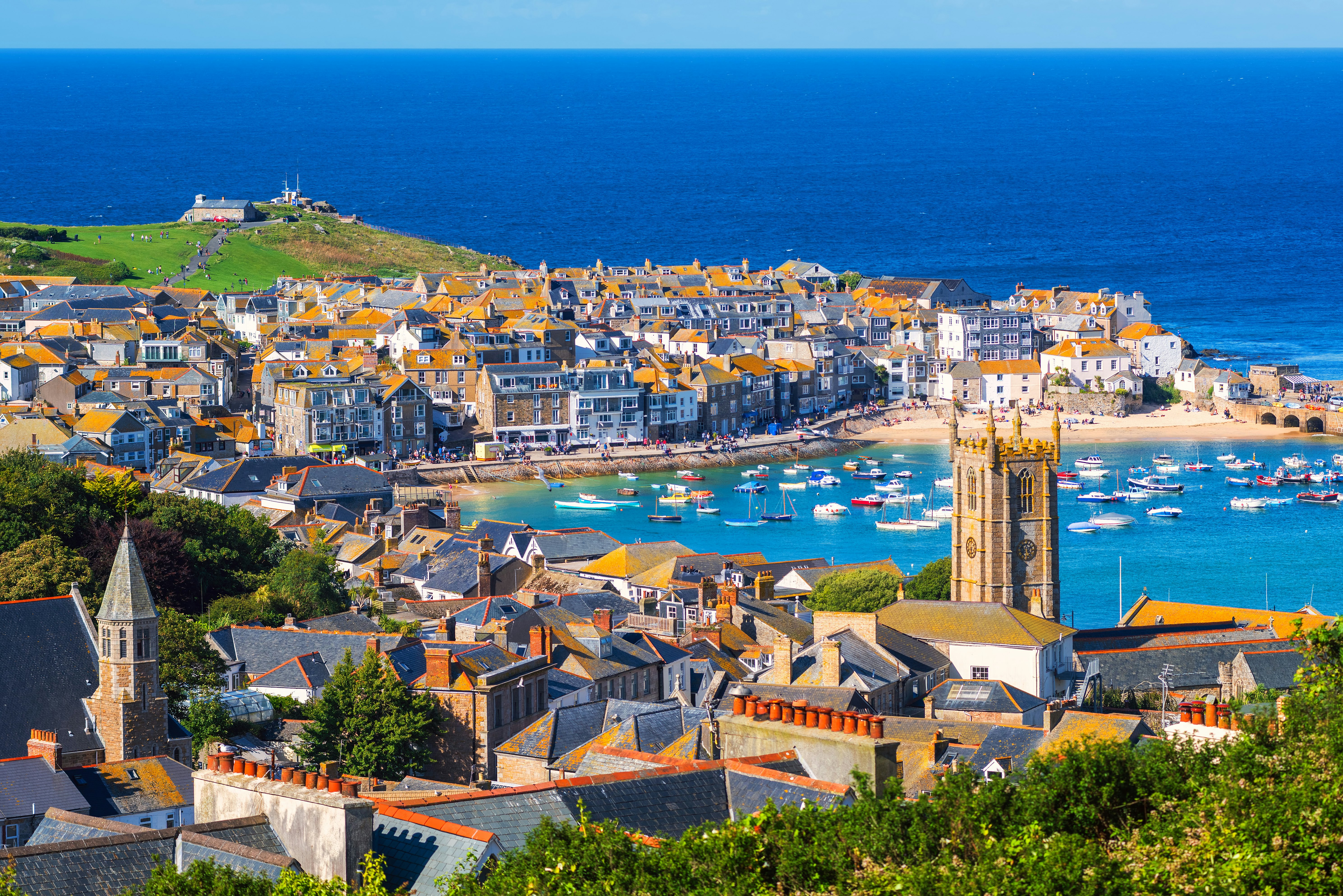Looking over the stone buildings and slate rooftops of St Ives, which curves round a harbour onto a grassy headland, with the bright blue Atlantic Ocean beyond.