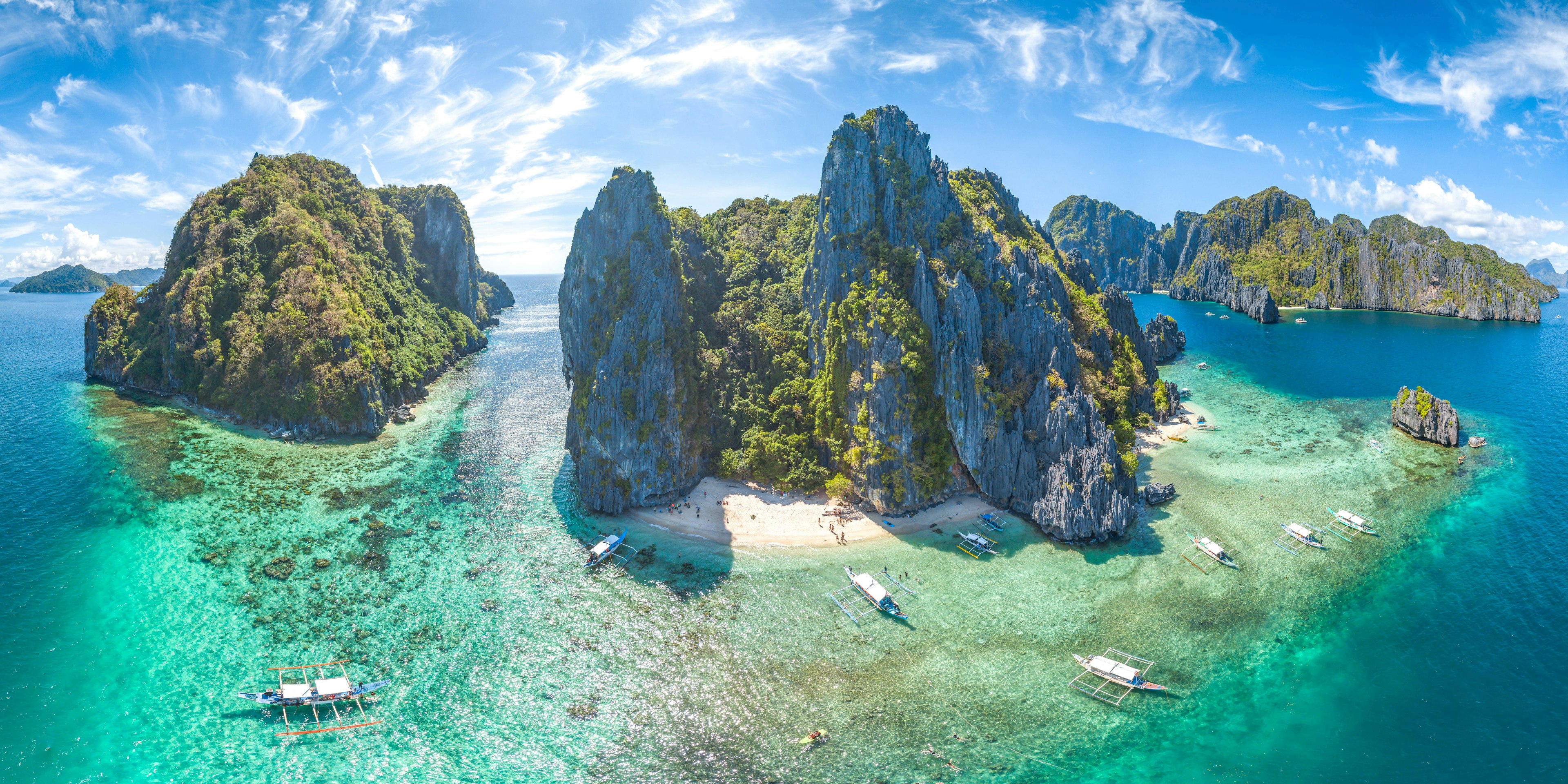 An aerial shot of El Nido, Palawan Province, Philippines. The small islands have very high rocky cliffs and lots of dense greenery. There are some white sandy beaches and a few boats moored near the islands.