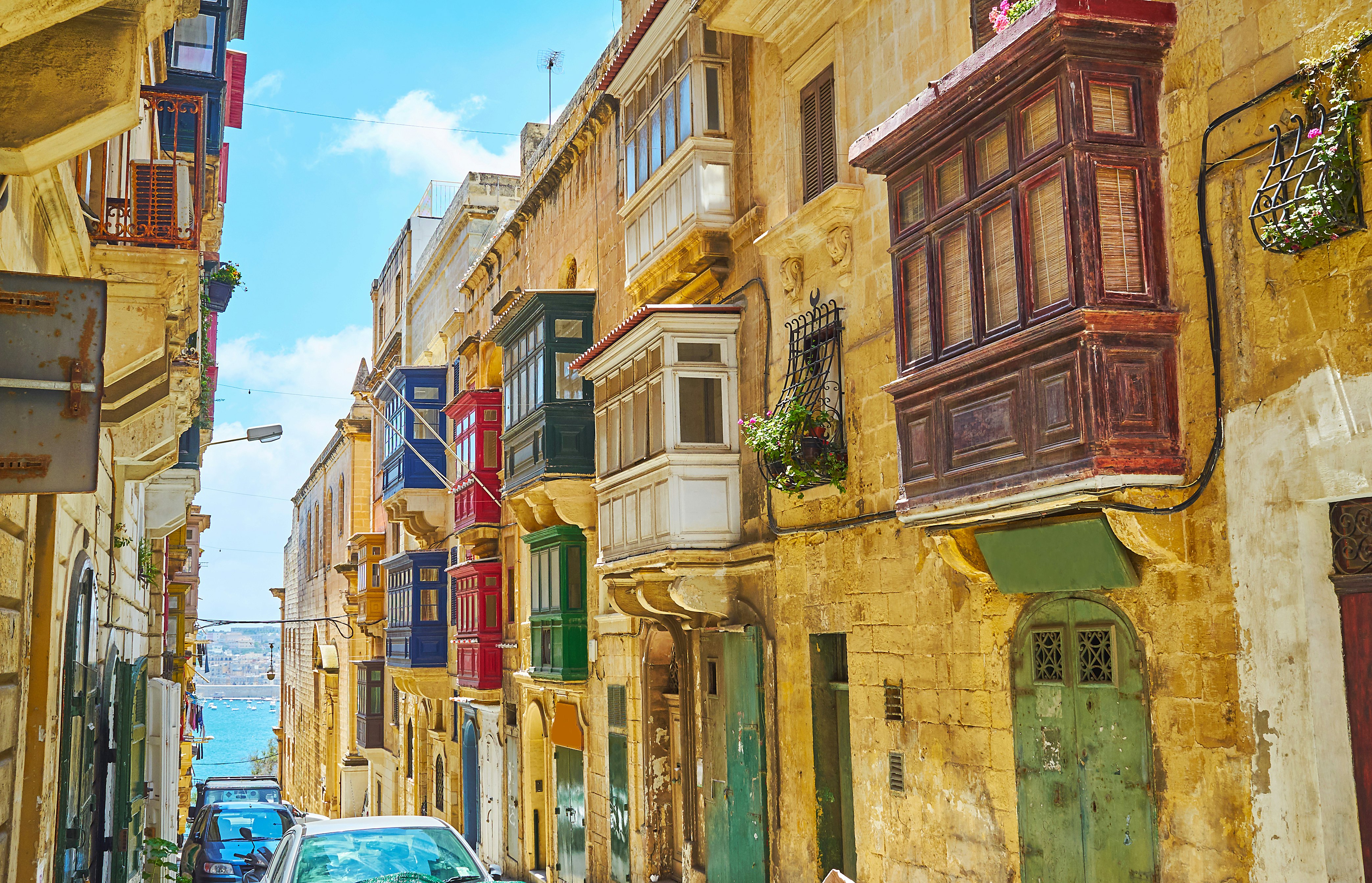 The narrow Archbishop street with parked cars and historical edifices with traditional wooden Maltese balconies. The blue waters of Grand Harbour is in the background.