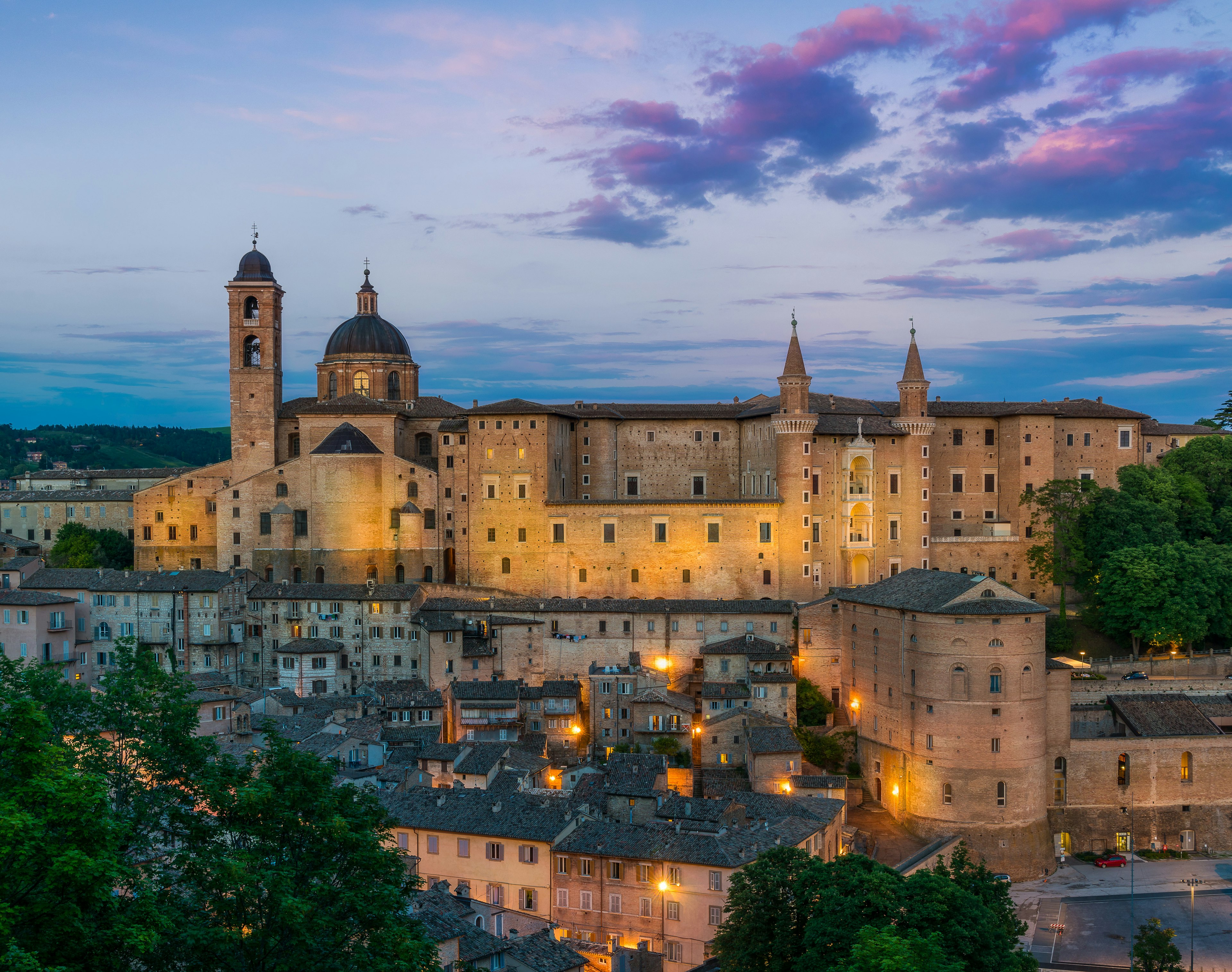 A view of Ducal Palace in Urbino at sunset; the sky is blue and purple and the palace is lit from below. Urbino, Le Marche, Italy.