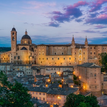 A view of Ducal Palace in Urbino at sunset; the sky is blue and purple and the palace is lit from below. Urbino, Le Marche, Italy.