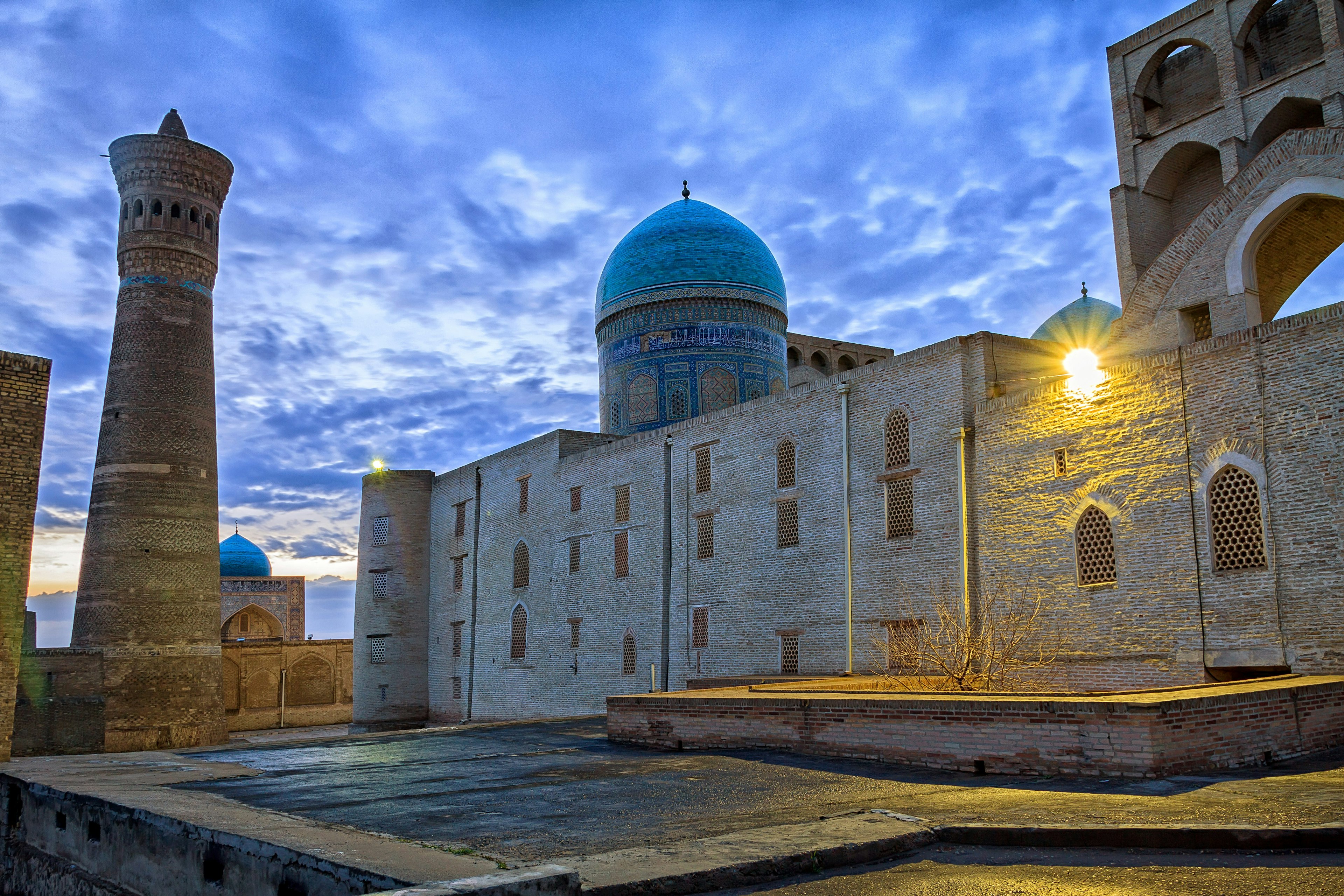 The blue dome and brown mud structures of the Kalon Mosque and minaret under cloudy, dusk skies.