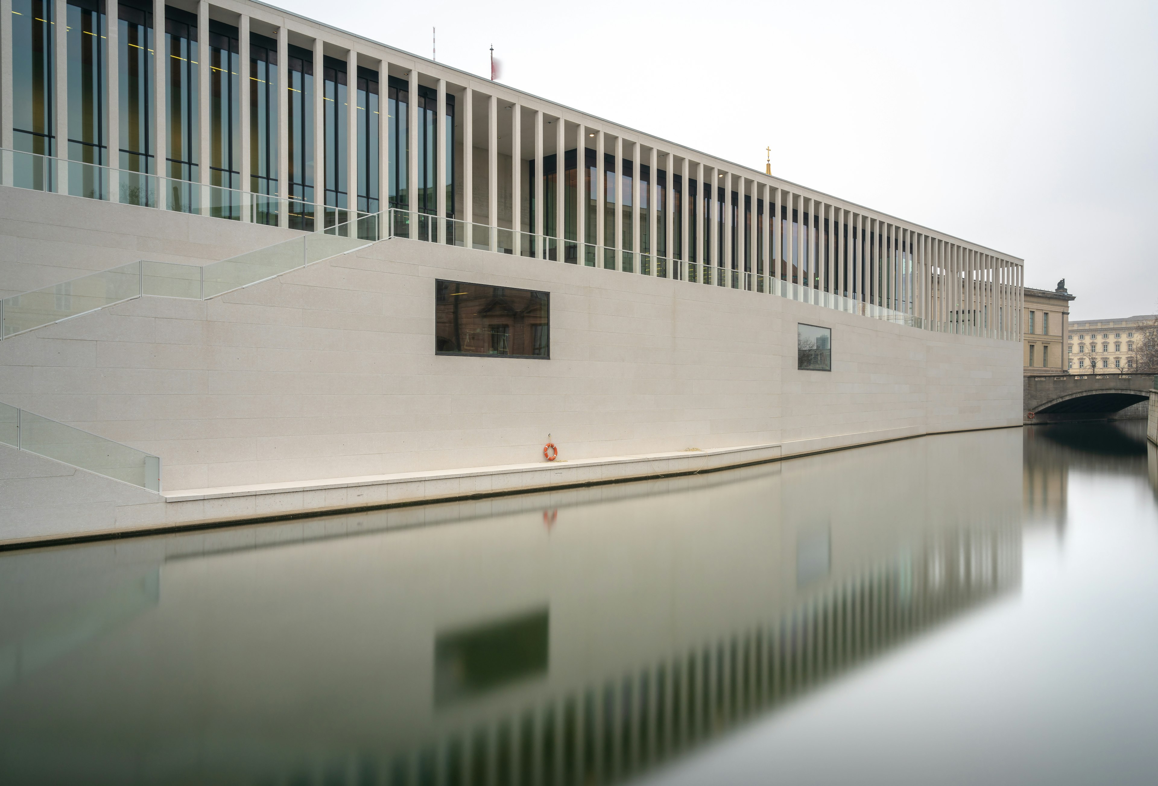 A photo of Museum Island's newest museum, the James-Simon-Galerie, taken from across the water. The building is very modern with large cream-beige bricks creating sleek lines. The windows are obscured by evenly spaced pillars/.