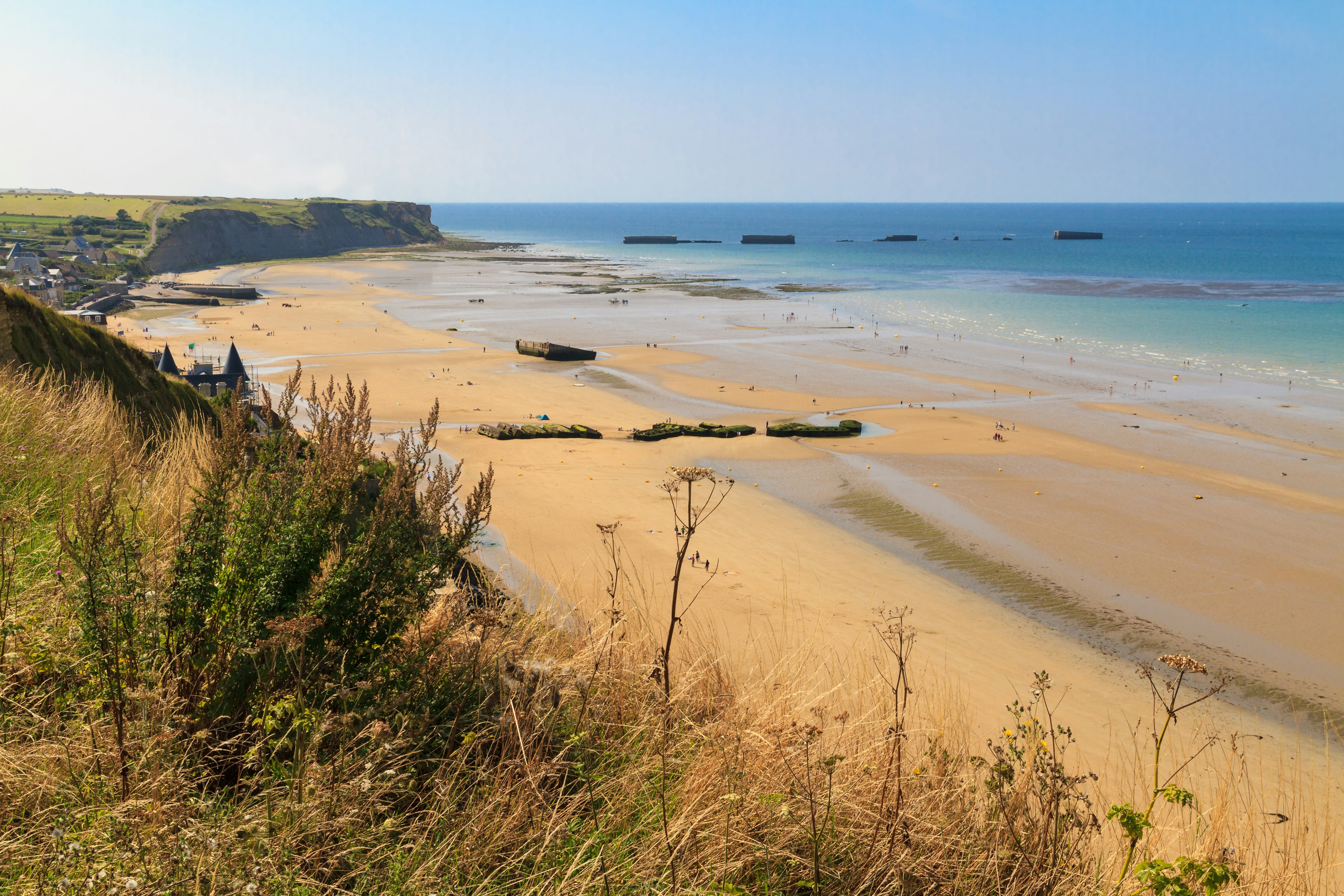 Looking down from a grassy clifftop over a huge golden D-Day landing beach, with the remains of a Mulberry Harbour (artificial port) at Arromanches-les-Bains visible in the distance