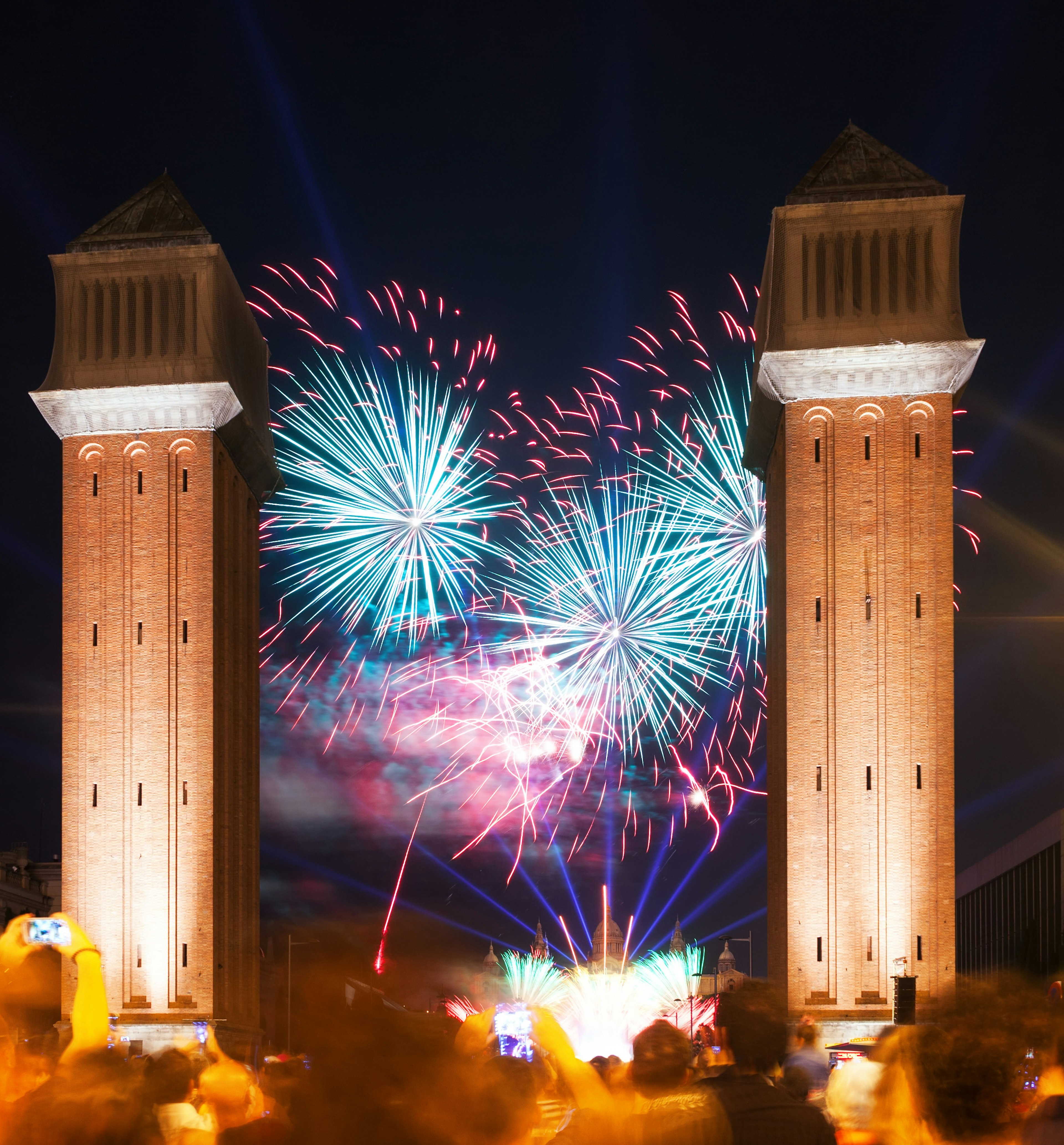 Blurred crowds watch a sky lit up with red and blue fireworks between two tall redbrick towers during a firework display at Plaça d’Espanya in Barcelona.