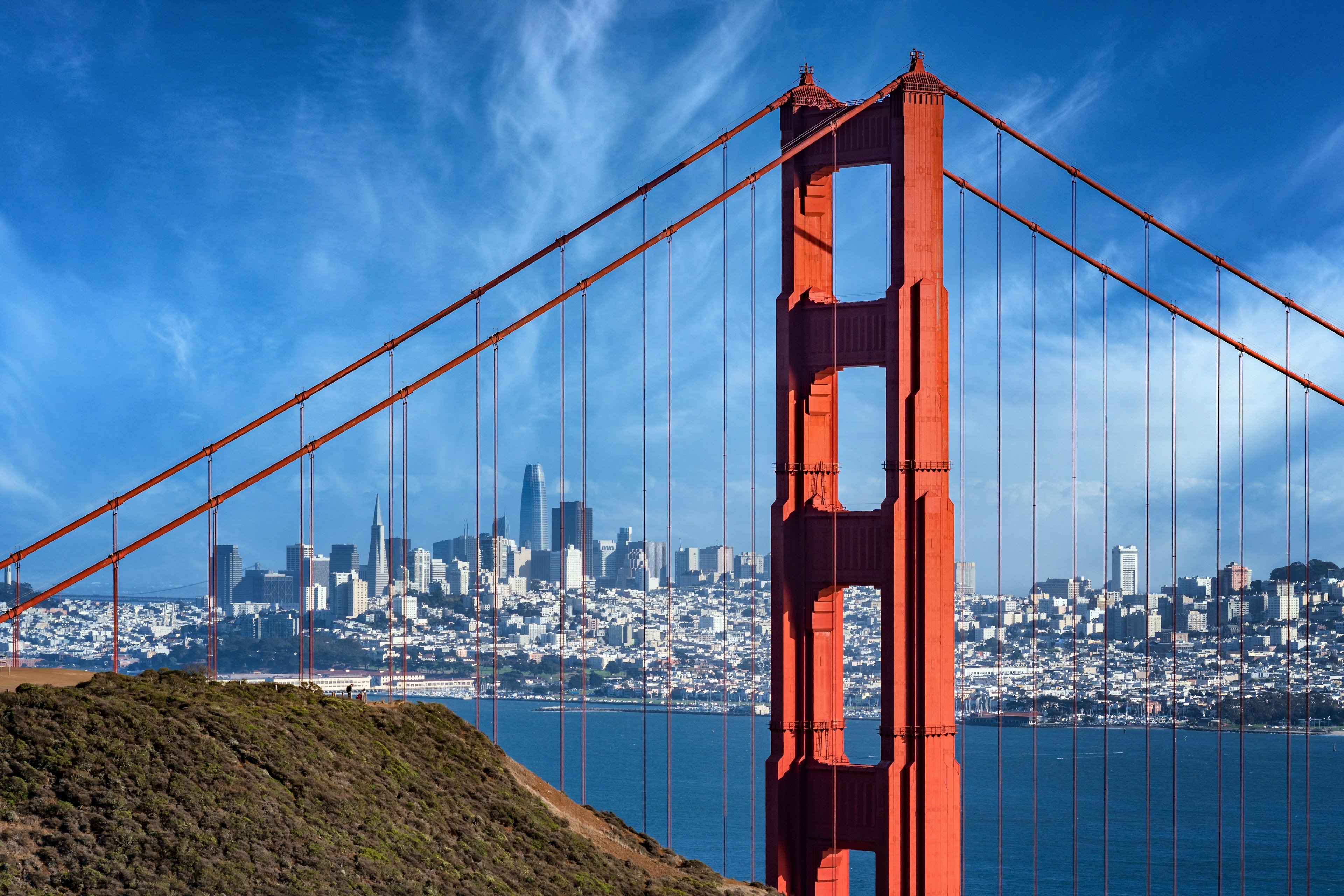 A large red-orange bridge stretches across a body of water. A high-rise city sits in the distance