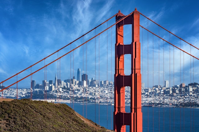 A large red-orange bridge stretches across a body of water. A high-rise city sits in the distance