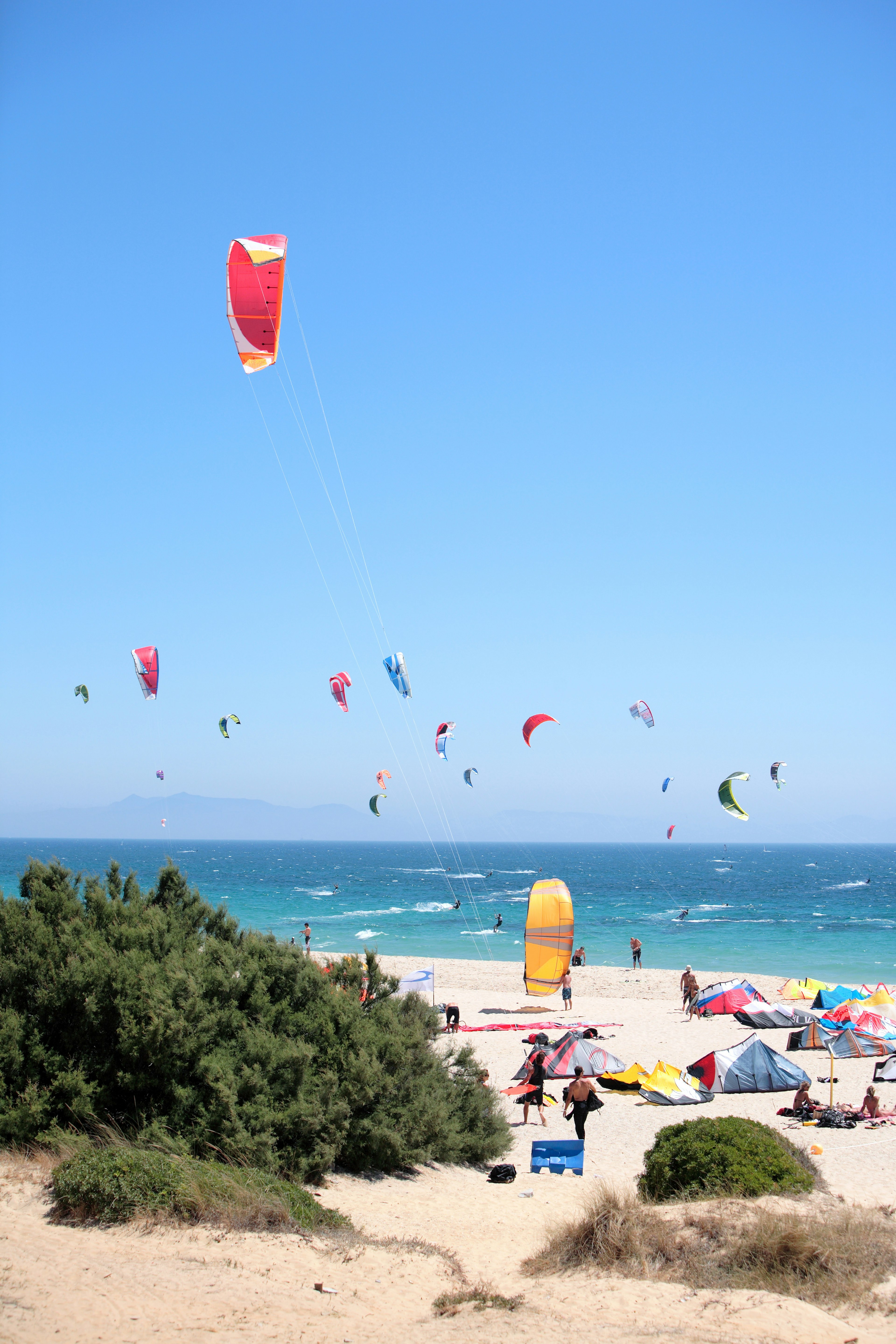 Tarifa beach packed with kite surfers and sunbathers, Colourful kites and tents stand out against the clear blue sky and white sand beach.