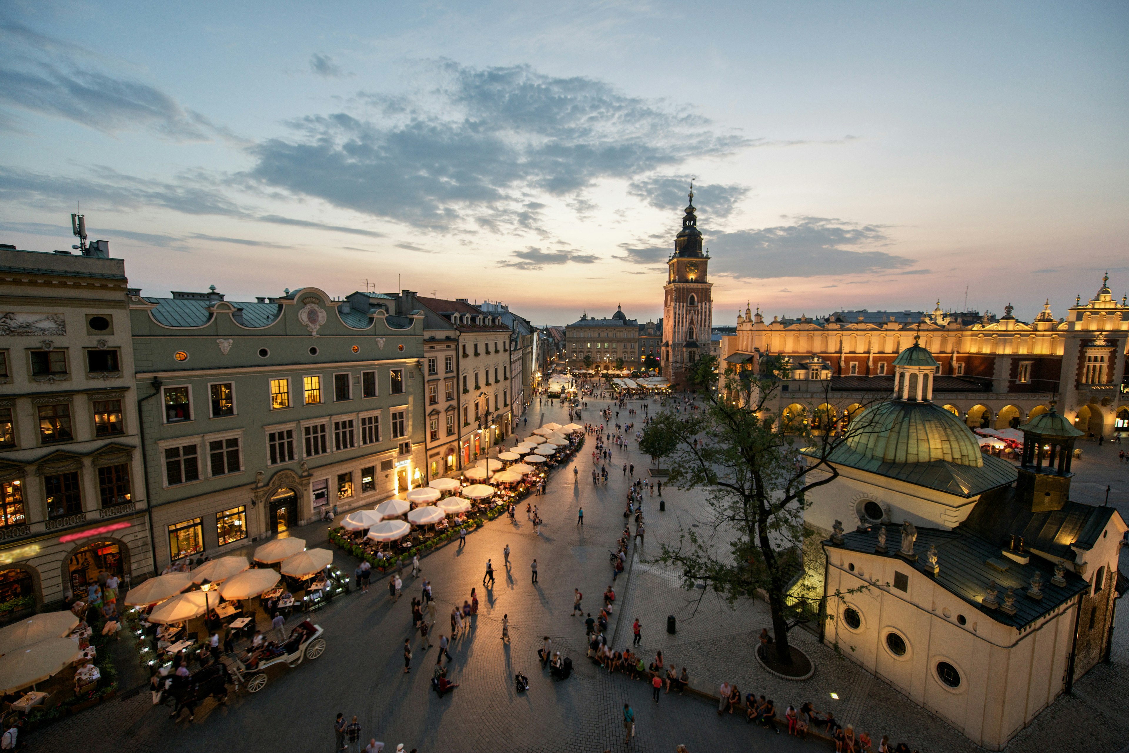 A view down over the square lined with grand buildings. The Town Hall Tower is the tallest of them all. Many outside tables at restaurants are occupied as the last light from the sun fades in the sky