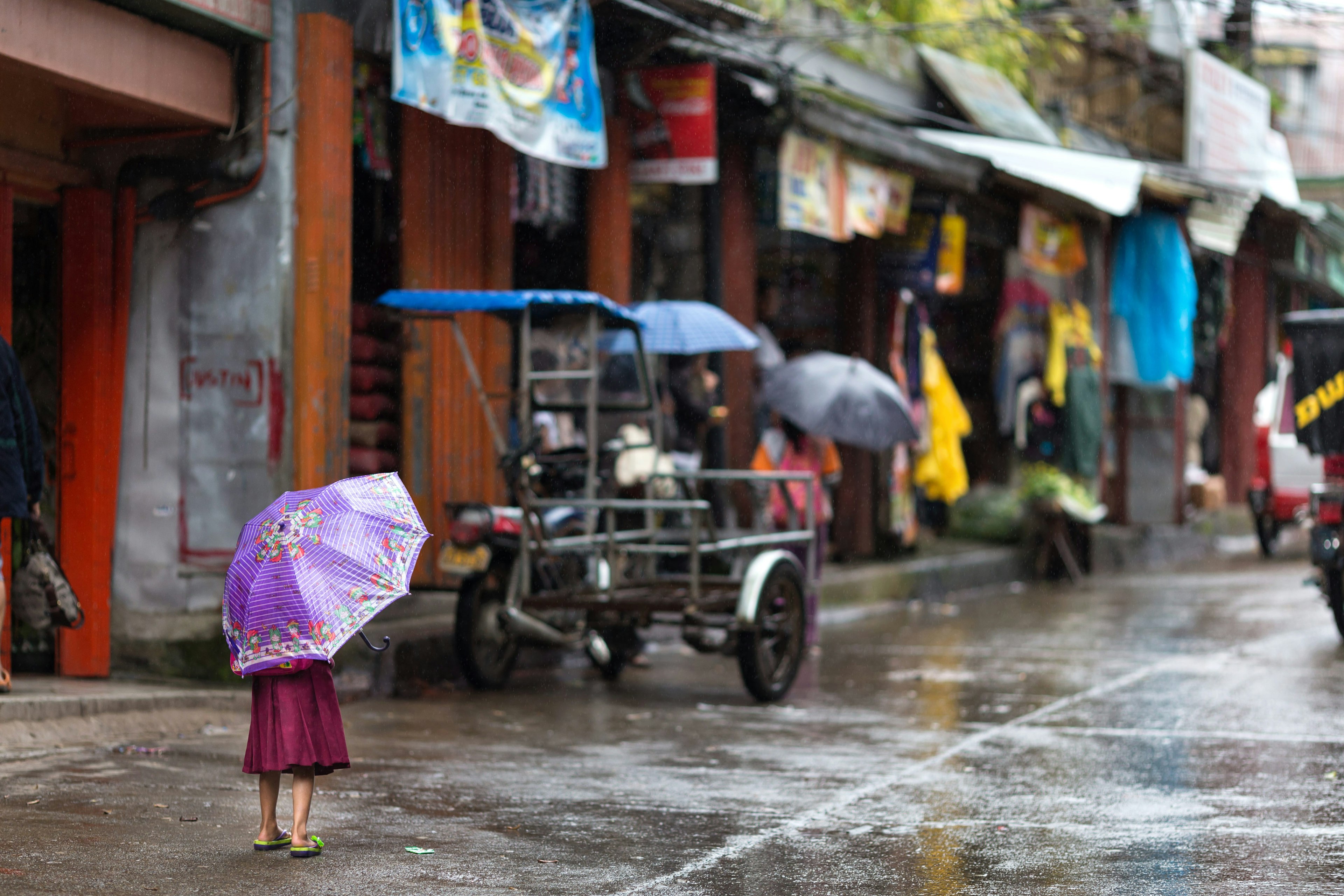 A little girl sheltering under a purple umbrella standing alone in the street in the rain in Banuae village, Philippines looking towards people sheltering in colorful shops