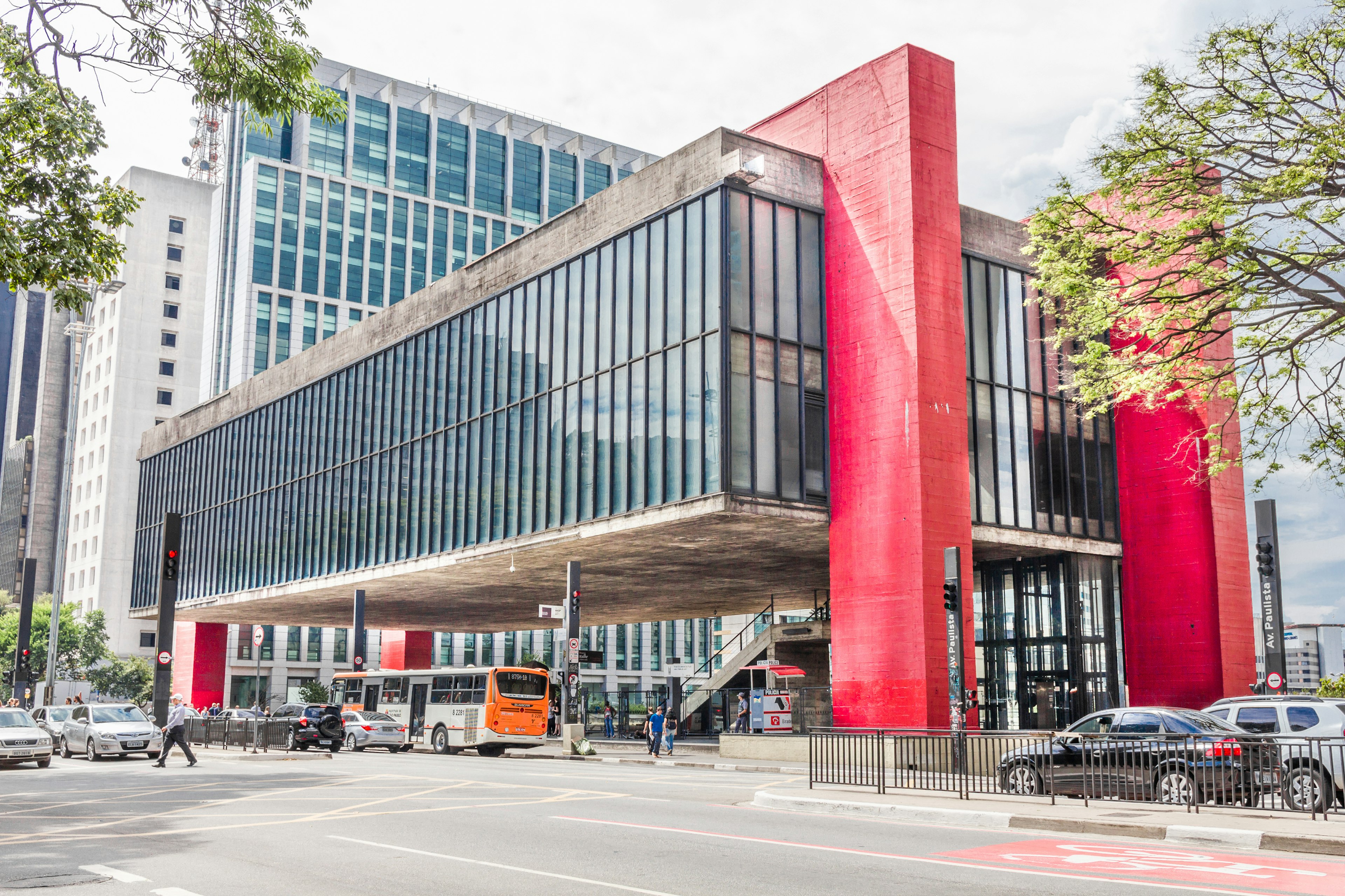 A concrete building with many windows raised from the ground on huge red blocks above a plaza busy with traffic.