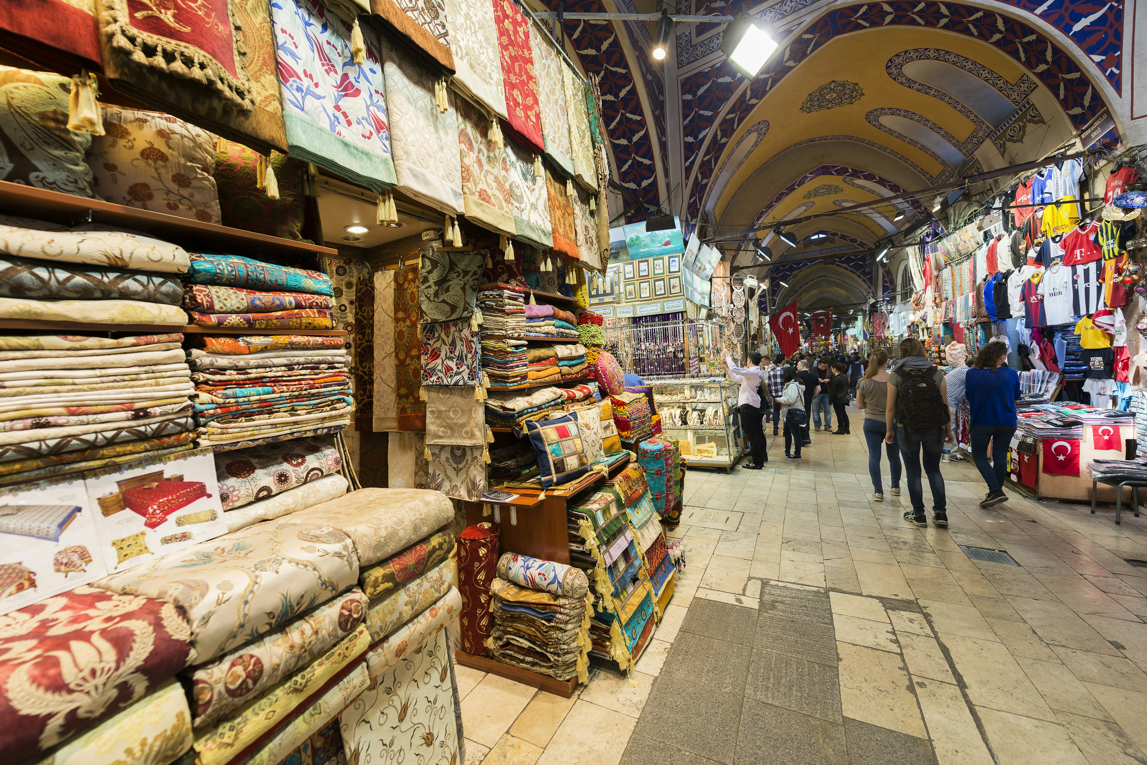 A display outside a shop within a large covered market shows many different heavily patterned textiles for sale.