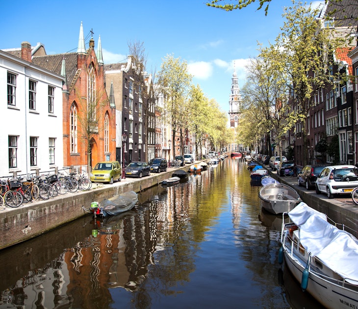 One of Amsterdam's central canals in the summer months. Boats full of people move through the water while the surrounding streets are thronged with people and bicycles.