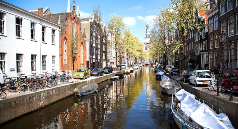 One of Amsterdam's central canals in the summer months. Boats full of people move through the water while the surrounding streets are thronged with people and bicycles.