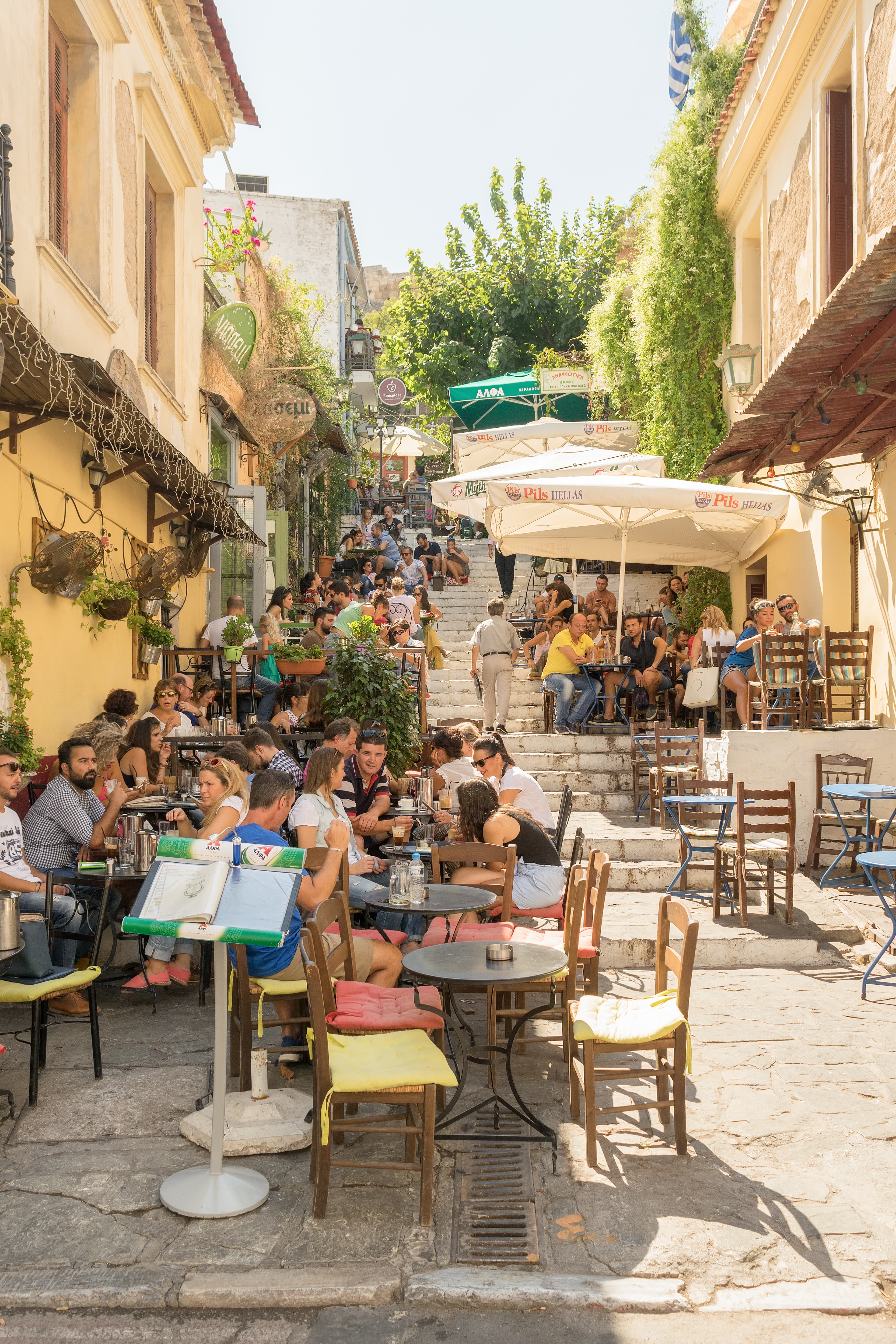 A pathway and series of steps are lined with tables and umbrellas. Many people are sat enjoying a drink