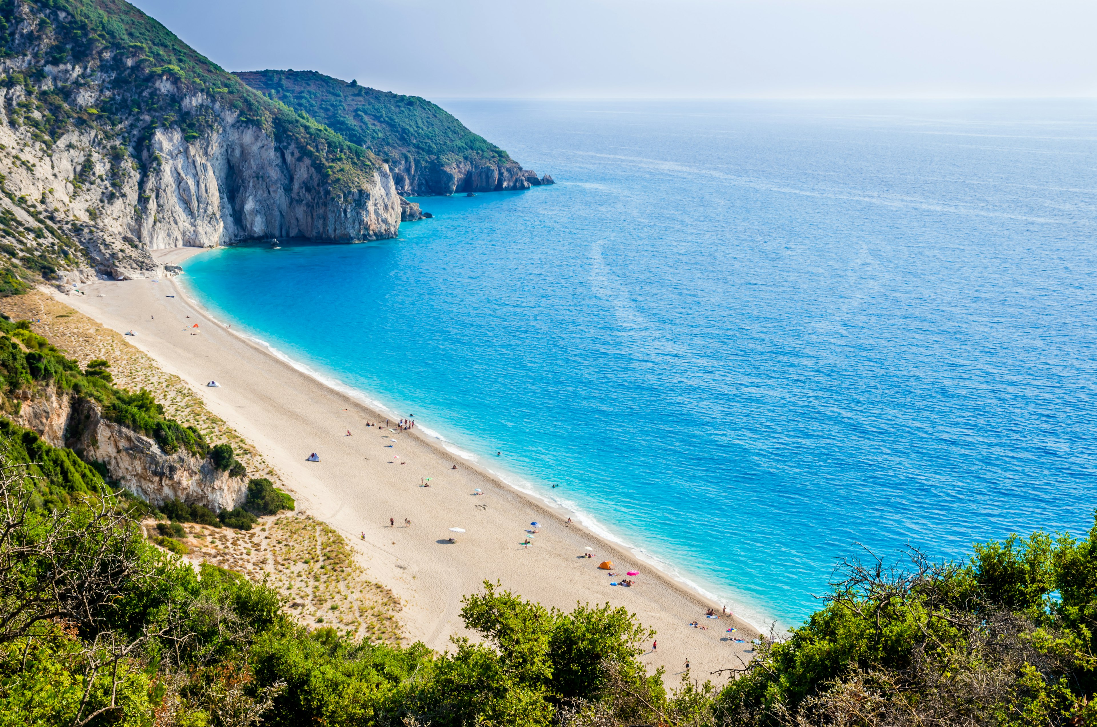 A view from the clifftop looking down towards a sandy beach and out to sea. There are a few colourful umbrellas and sun tents down on the beach