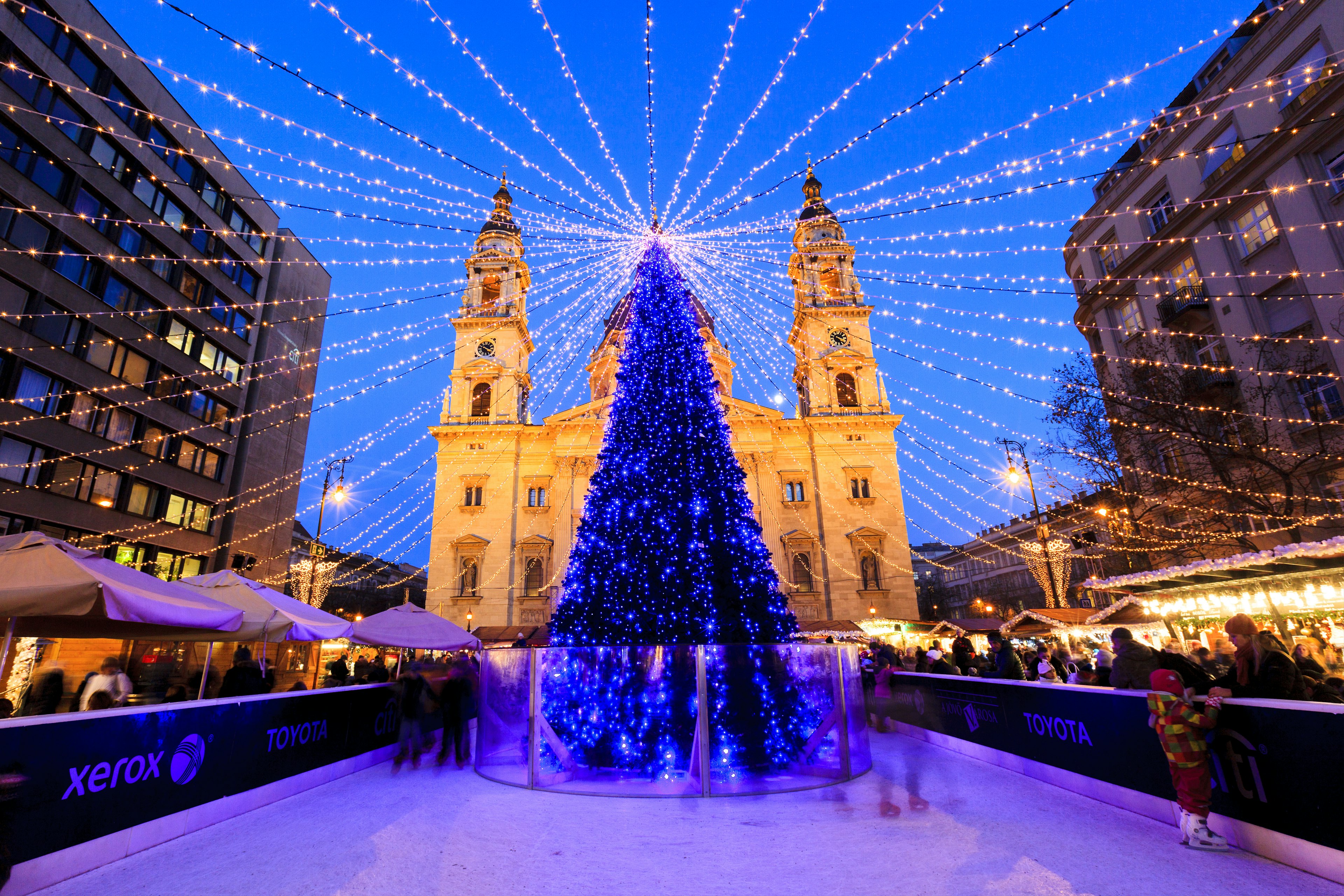 A tall Christmas tree covered in small blue lights stands in the middle of an ice rink in front a large church. Strings of lights fan out from the top of the tree to form a canopy of lights above the ice skaters