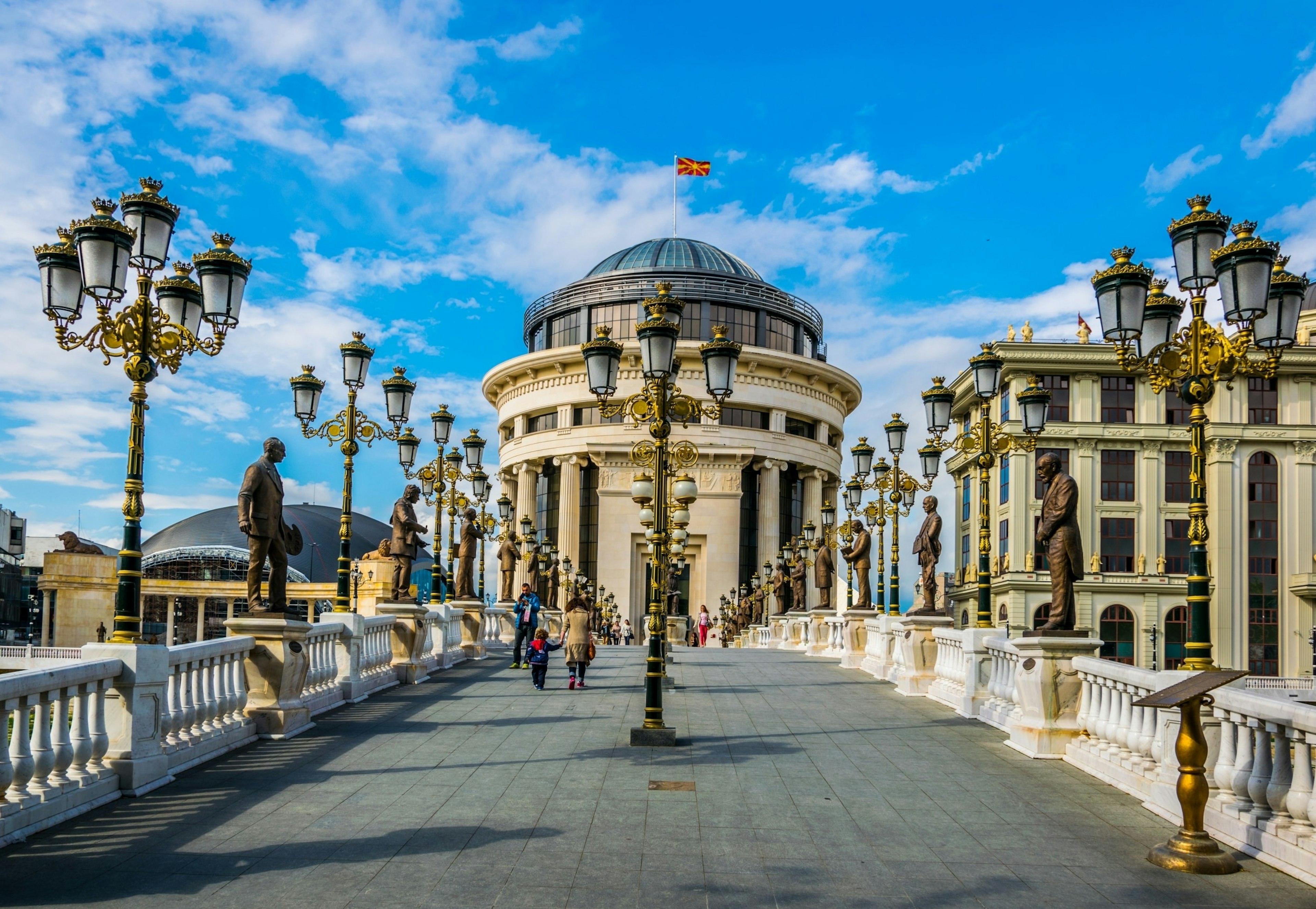 A row of ornate golden street lamps line the edges of a bridge leading to a pillar structure; North Macedonia Best in Travel