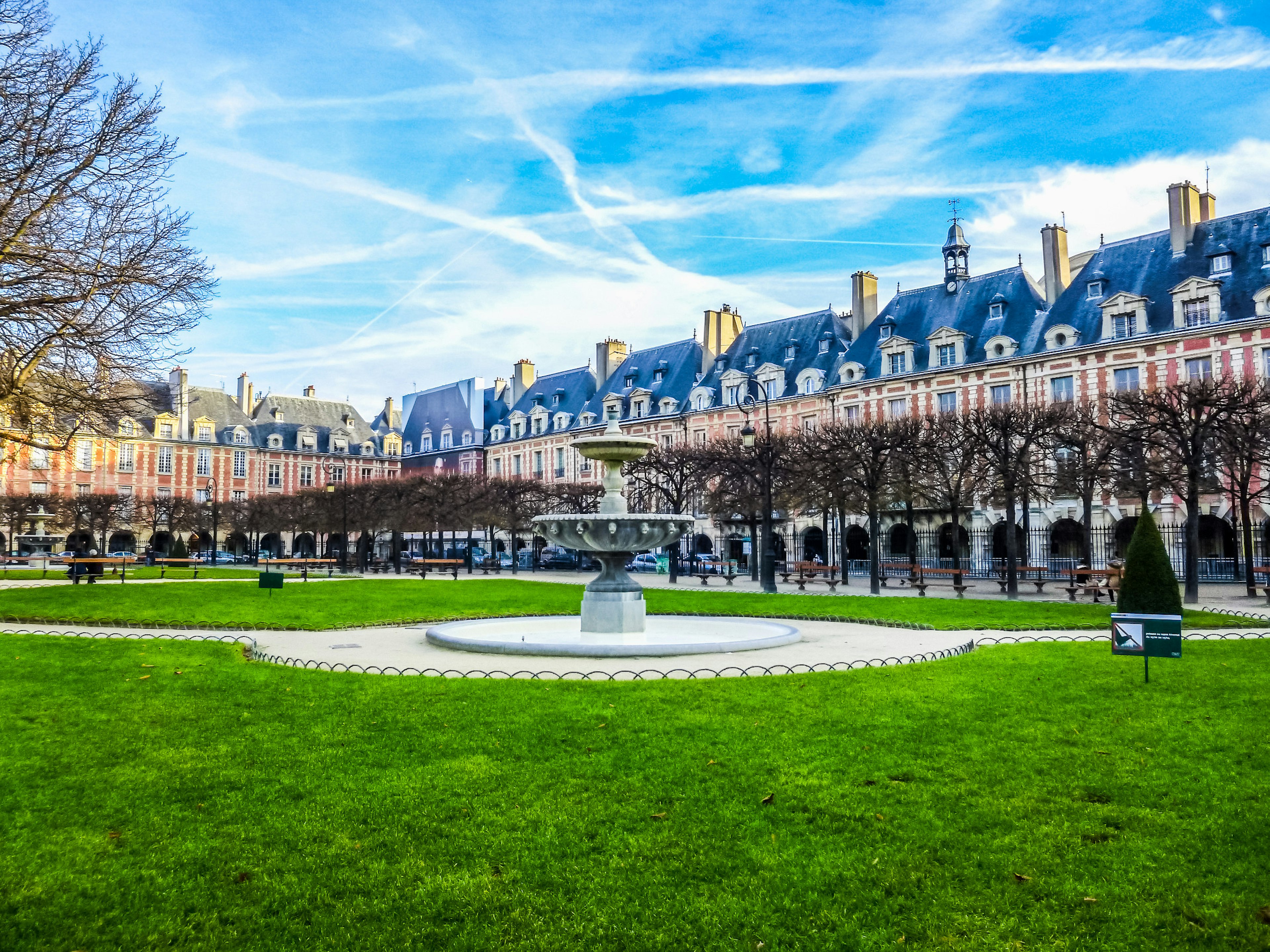 A view of sunny Place des Vosges, a patch of green park surrounded by traditional townhouses, in Paris.