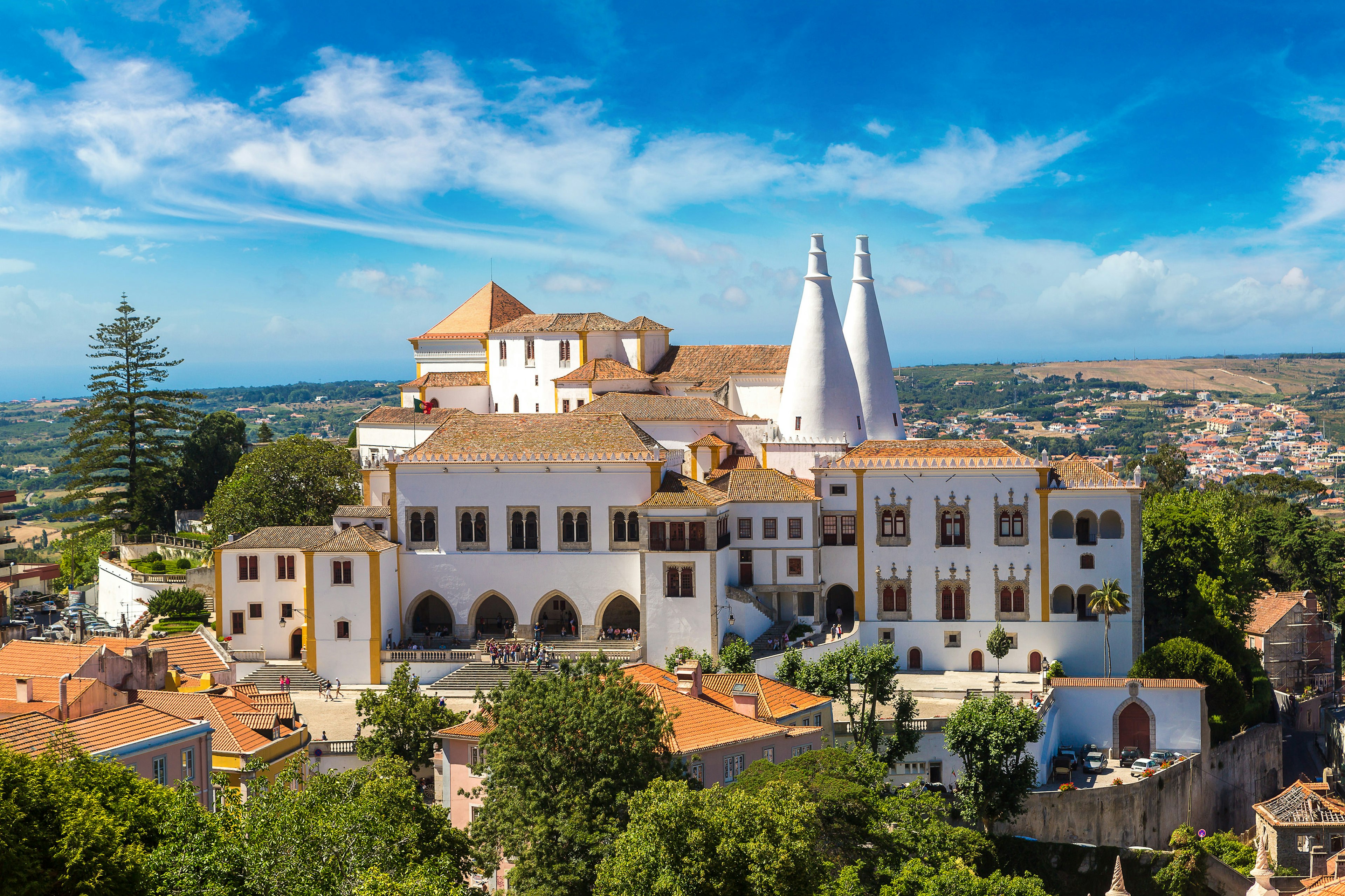 Photo of Palácio Nacional de Sintra (Sintra National Palace); the large complex sits on top of a hill and has whitewashed walls, a terracotta-tiled roof, and two conical white turrets.