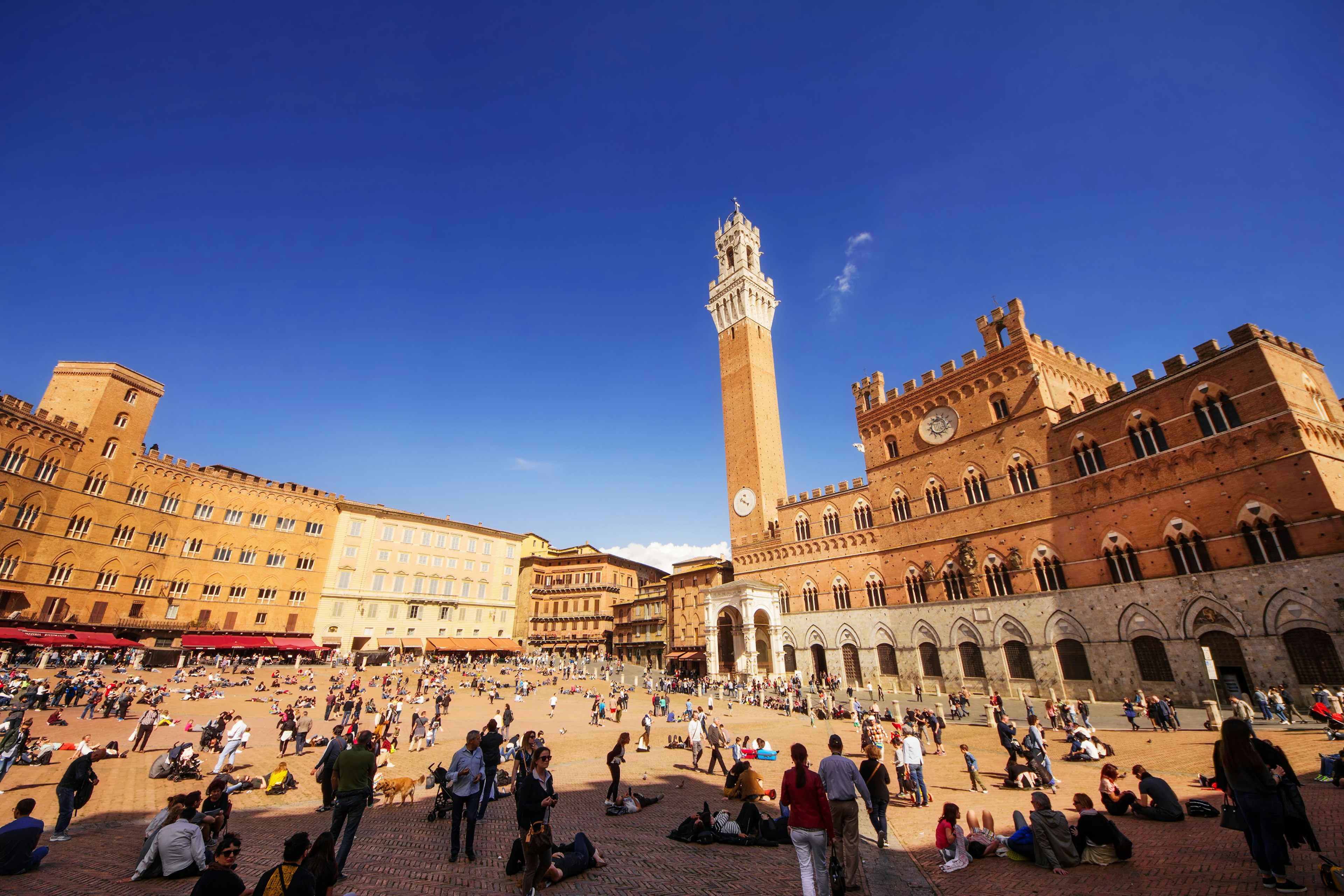 A medieval main square in a small city. People are gathered in groups in the sunshine. A tall bell tower looms over the square