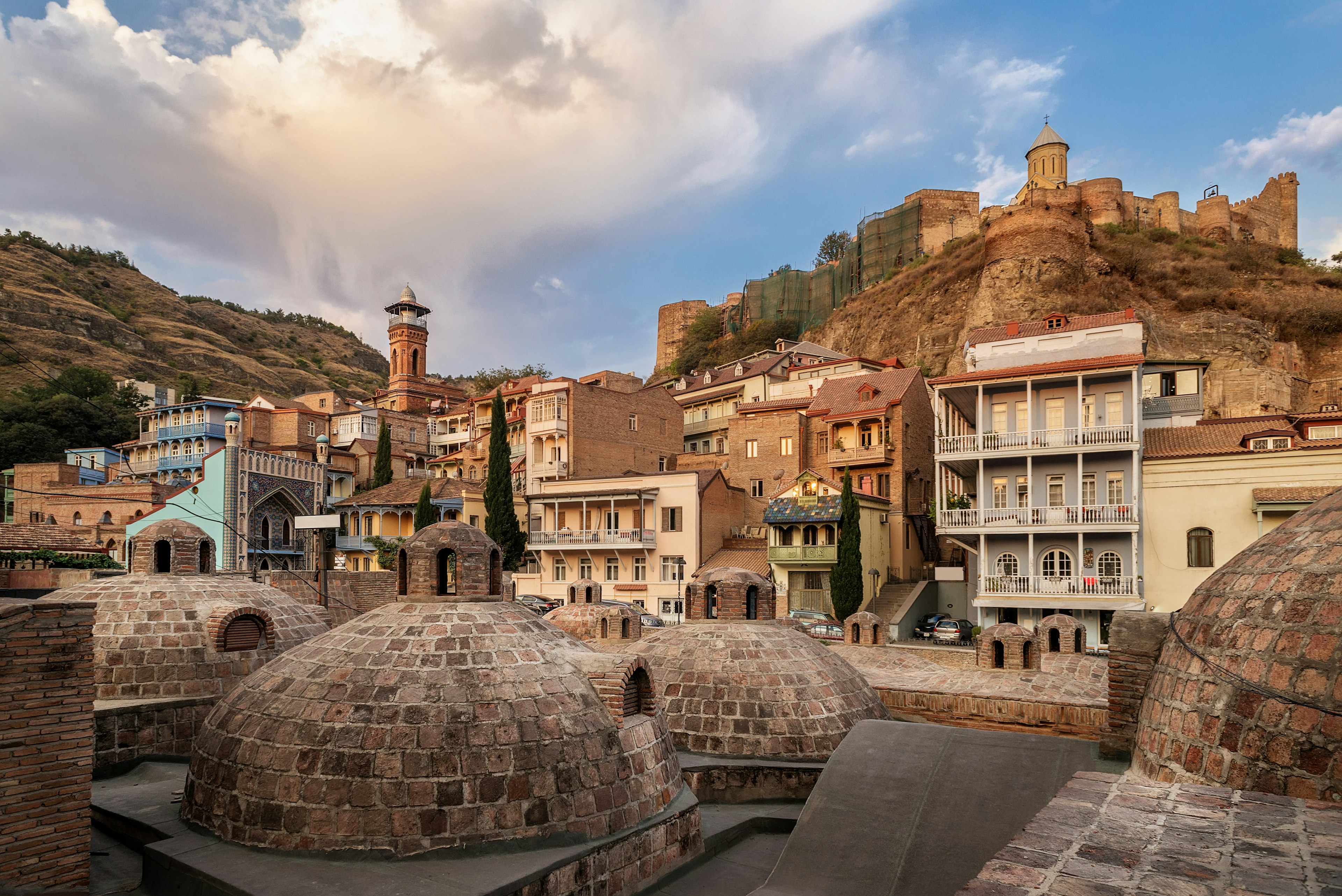 A series of brick domes on a rooftop. Above them are homes built into the side of a hill, with a fortress at the top