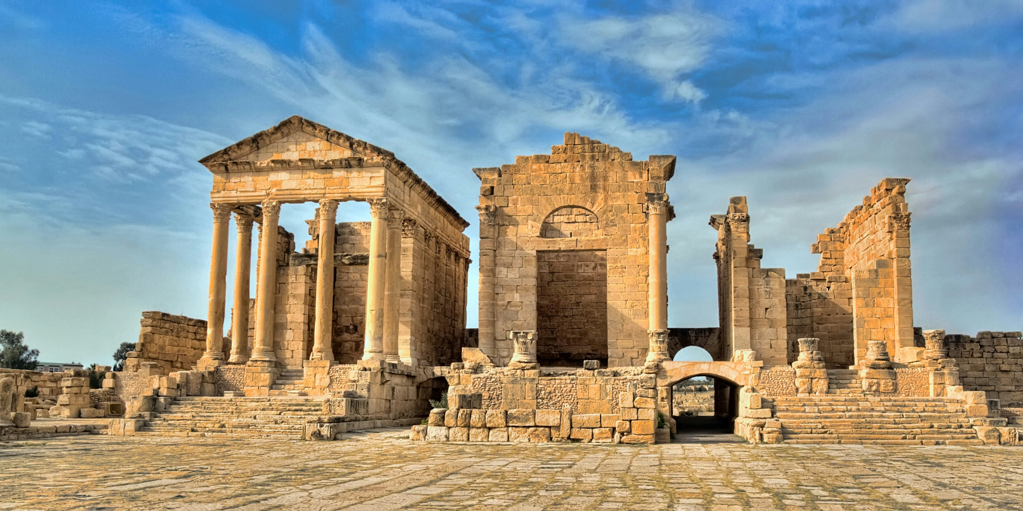 Three large temples rise up into a blue sky; the one on the left is in the best state of repair, with its facade's columns still holding up its triangular roof; the one in the middle has lost most of its columns and upper facade, while the one on the right is only a skeleton of the lower walls.