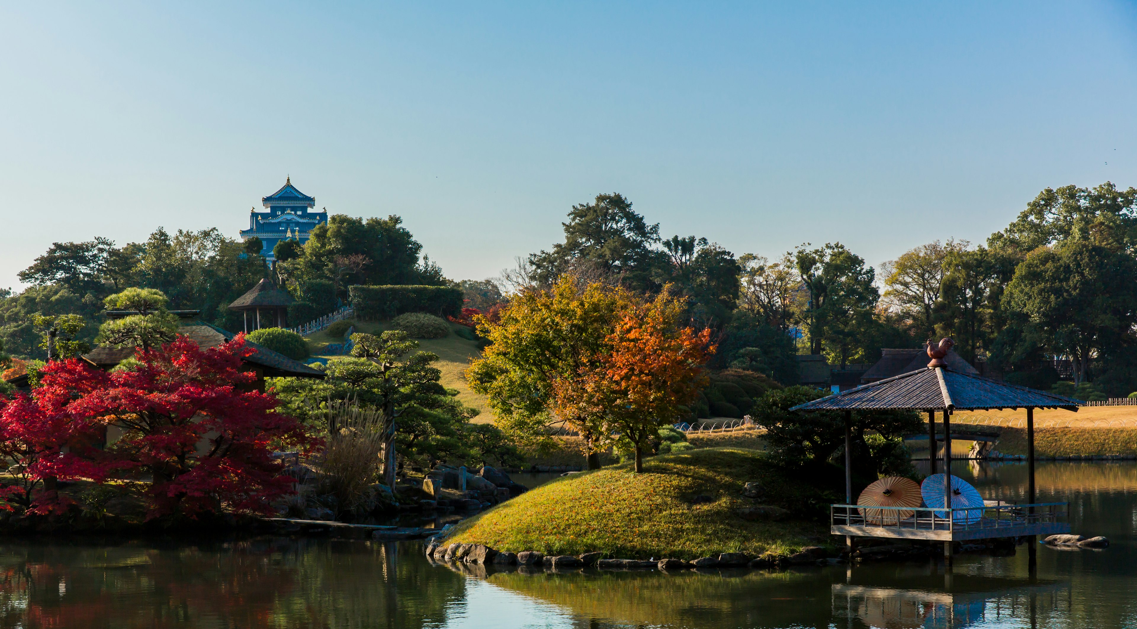 A view of Koraku-en garden in Okayama, with the still lake in the foreground leading away to green hills dotted with trees and an ornate castle visible in the background.