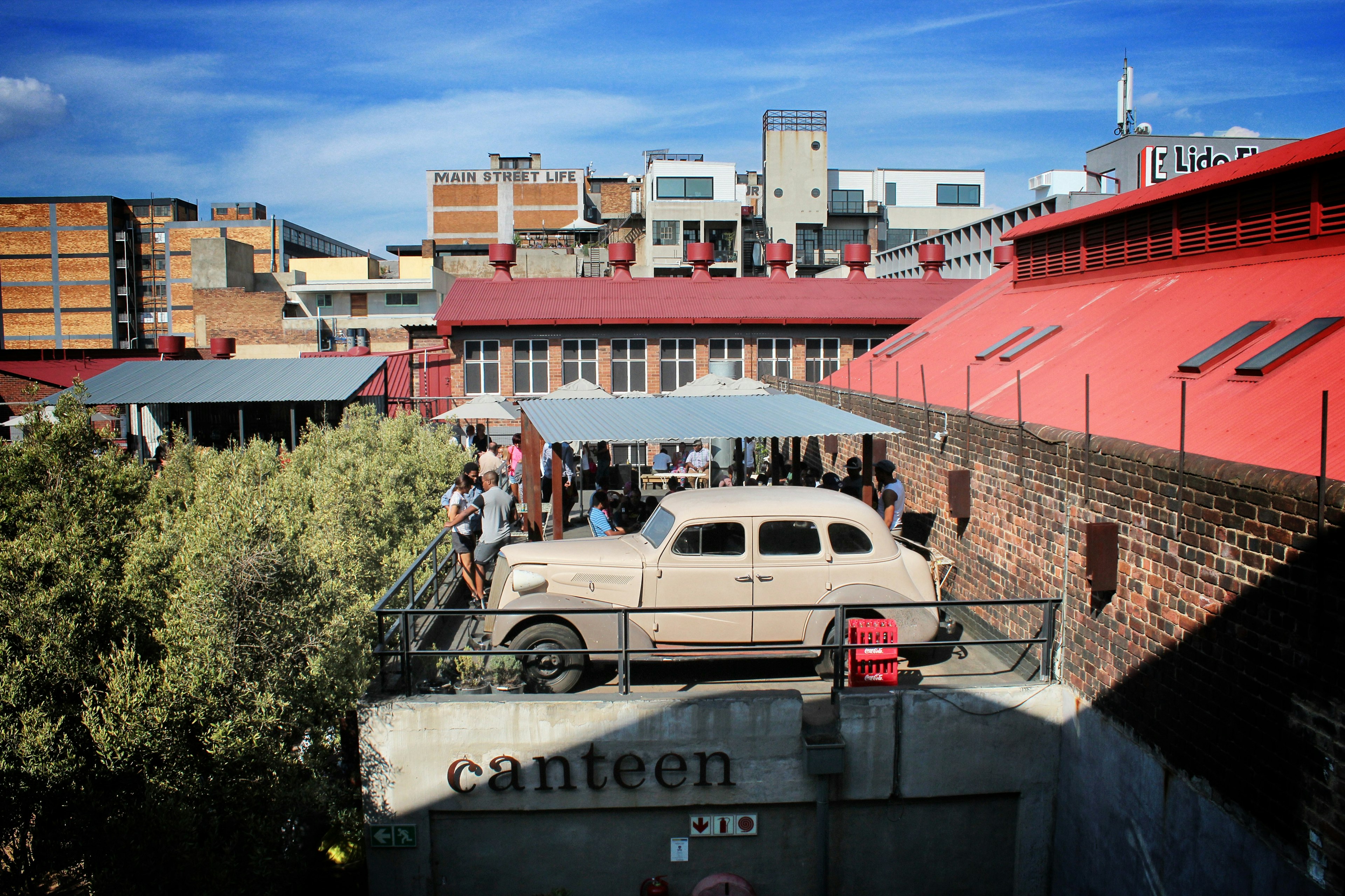 A crowd gathers under an awning on the flat roof of Market on Main, sharing the space is a vintage 1930s car. Trees climb up to this level on the left and renovated old industrial buildings back the scene.