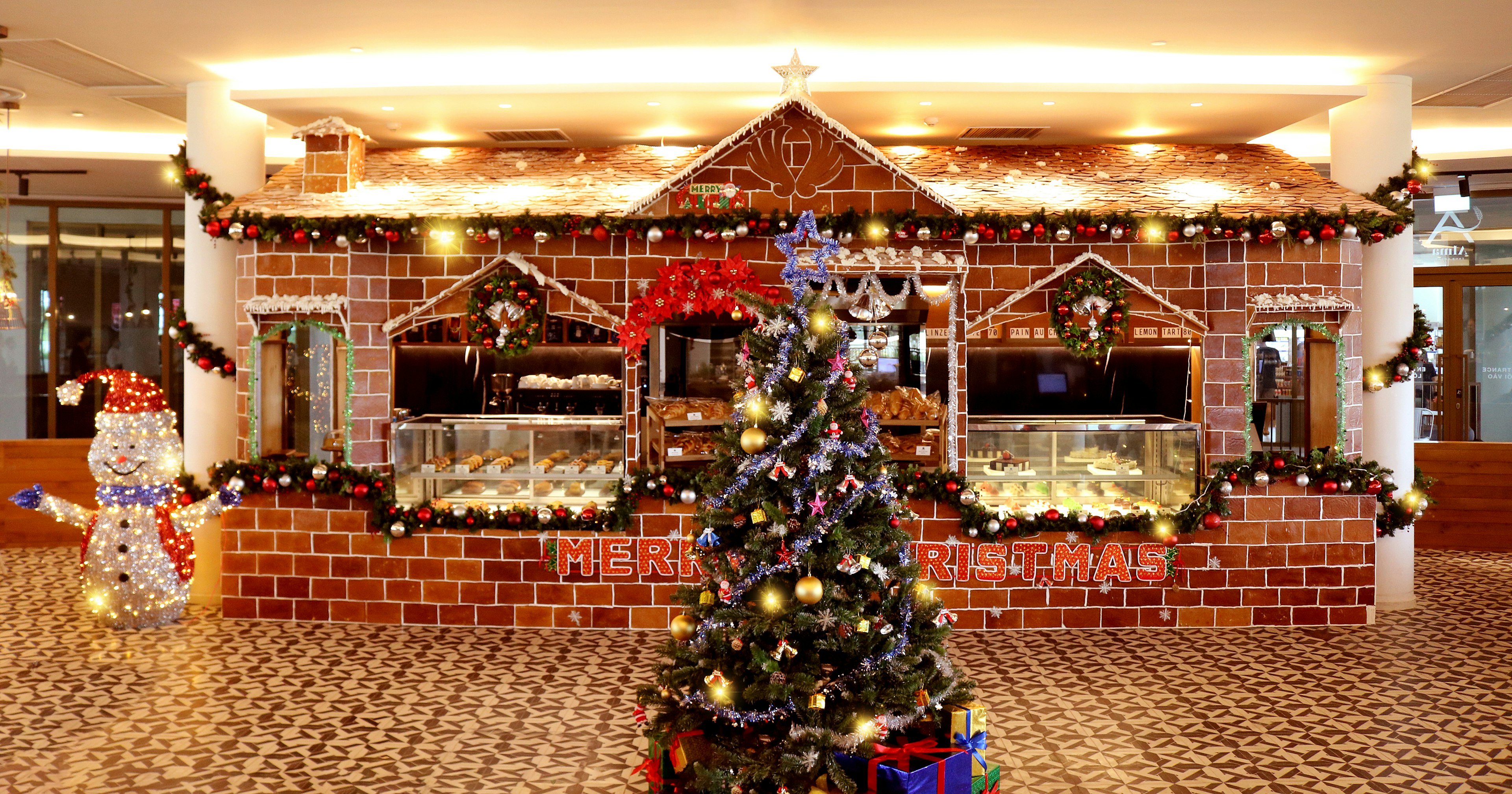 A Christmas tree in front of a large gingerbread house surrounded by tinsel and a snowman model