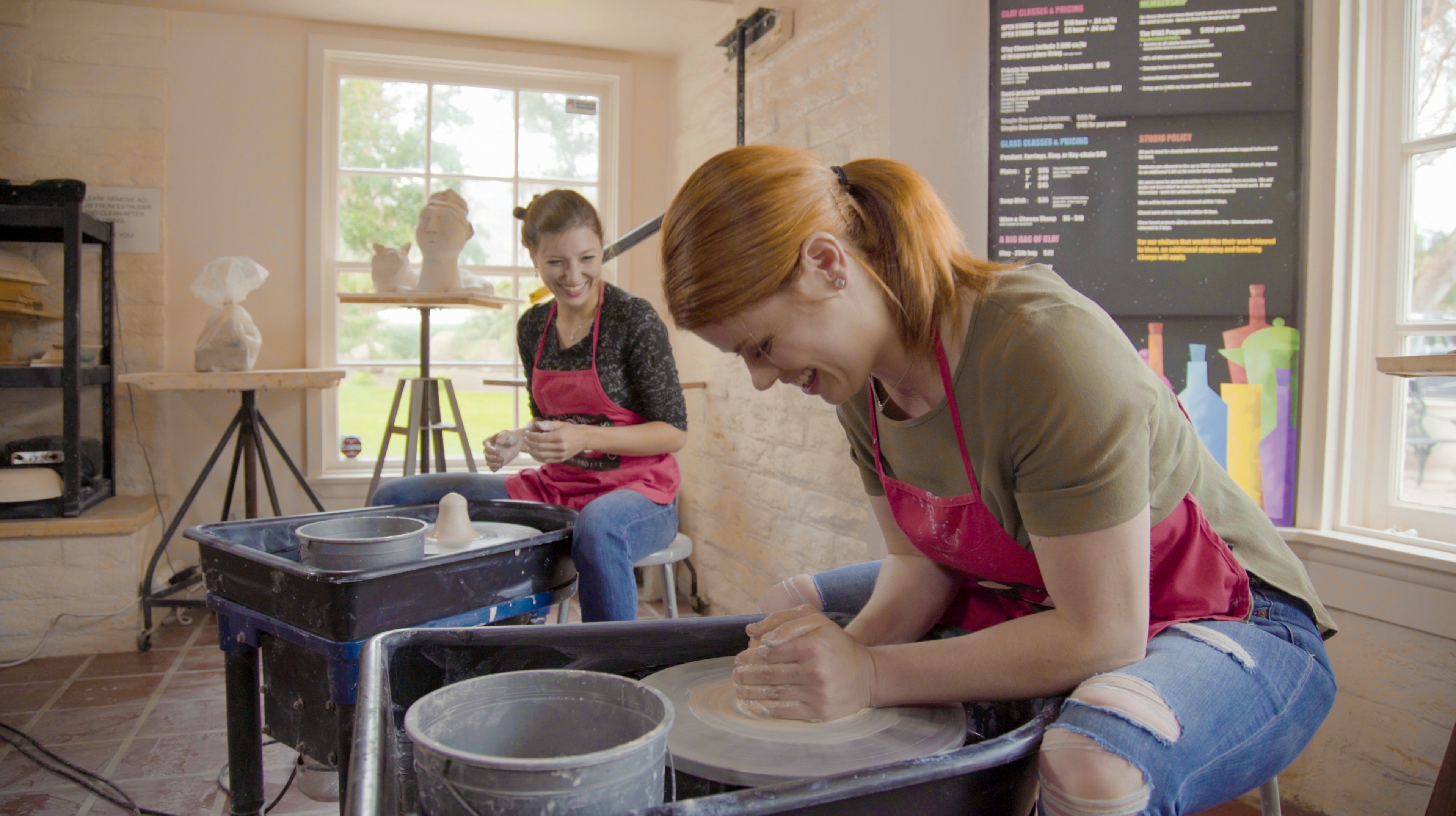 Two women use potters wheels to make ceramic artworks while wearing red aprons at a gallery and workshop in Greater Palm Springs