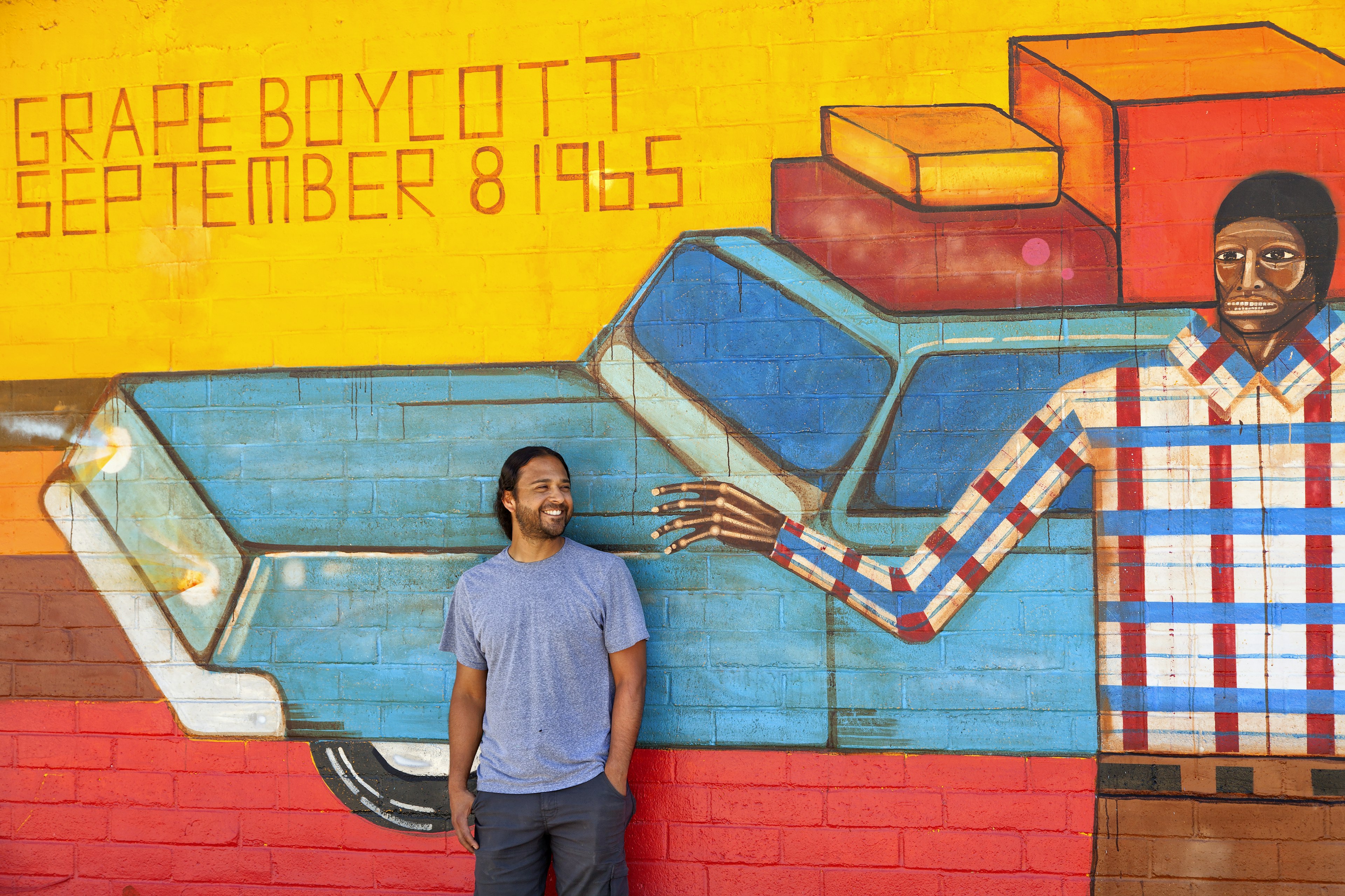 An artist stands in front of a wall mural that fills the frame. The mural is bright yellow and red, with a bright blue station wagon in the center and a black man in a checked shirt.