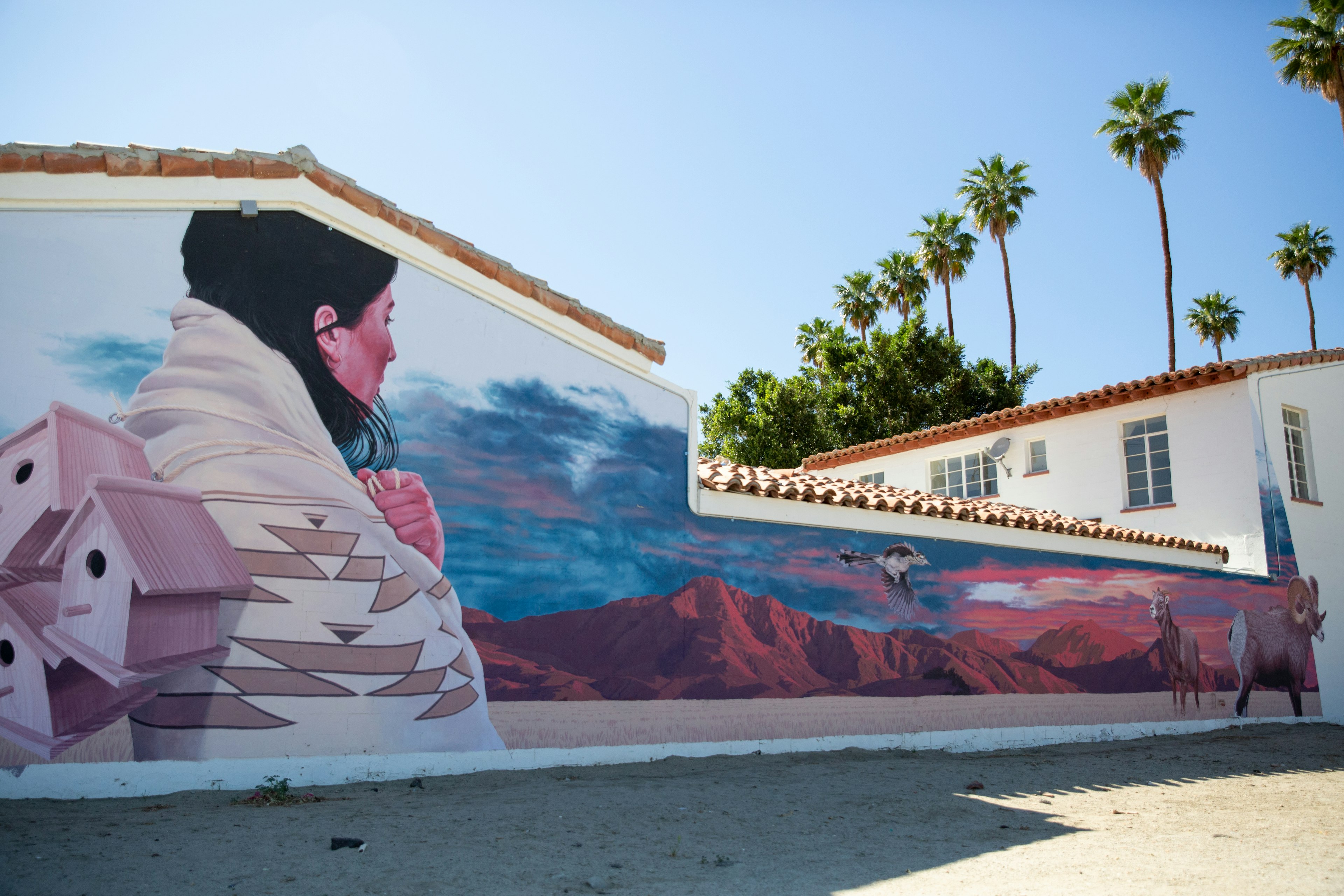 A mural on the side of a house shows a Native American woman wrapped in a patterned blanket and carrying three wooden birdhouses toward red hills in the sunset.