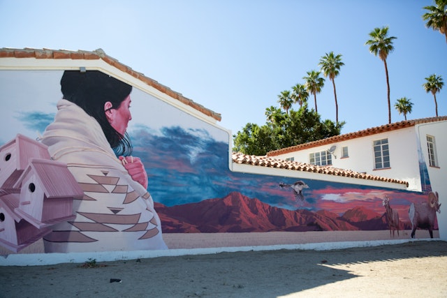 A mural on the side of a house shows a Native American woman wrapped in a patterned blanket and carrying three wooden birdhouses toward red hills in the sunset.