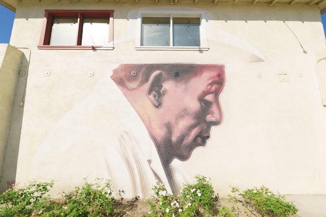 A painted mural on a wall shows a hispanic man in typical farm working clothing, with a brimmed hat, as he gazes downward and to the right