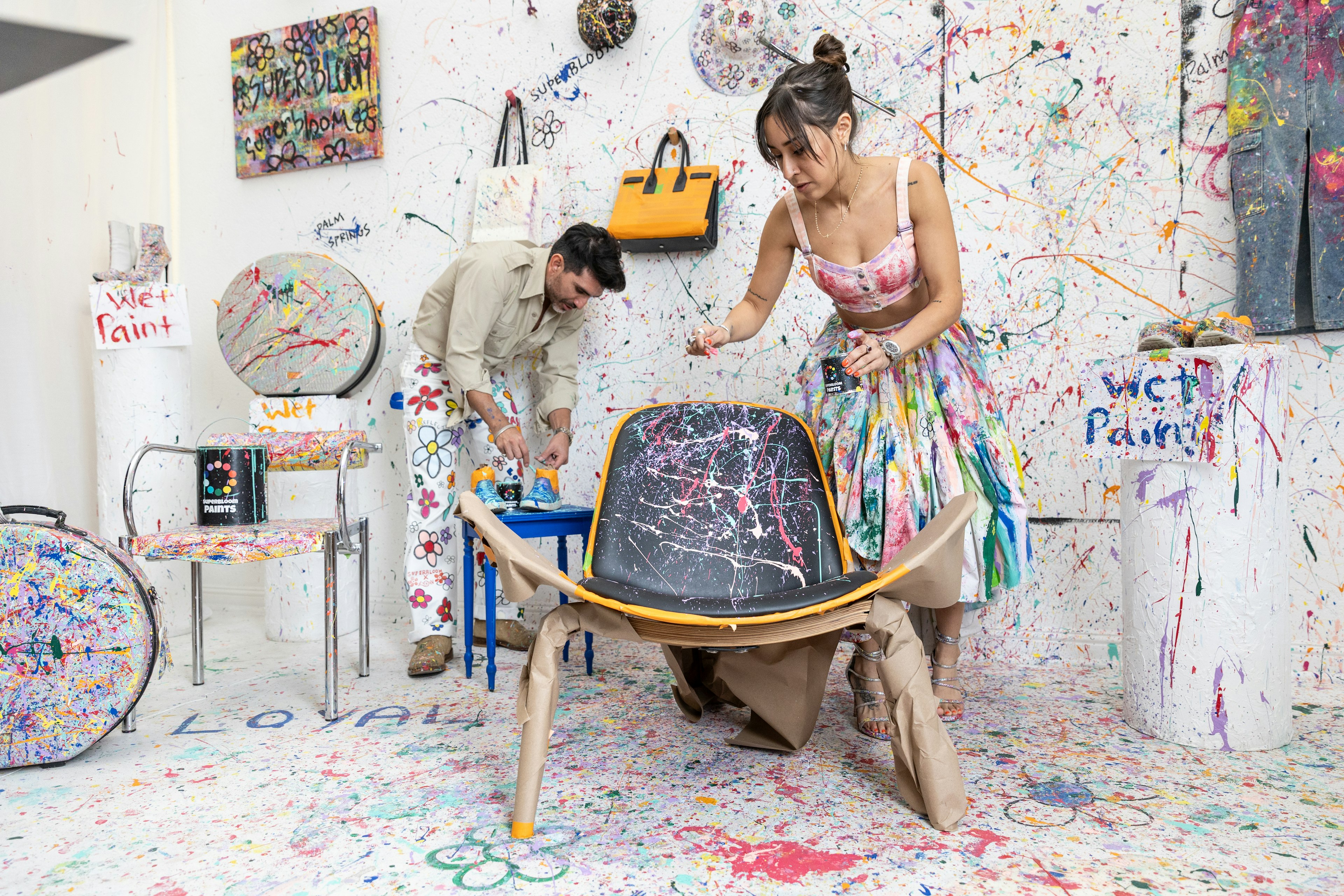 Two people splatter multicolored paints over a midcentury modern chair as spattered paint covers the walls and other objects