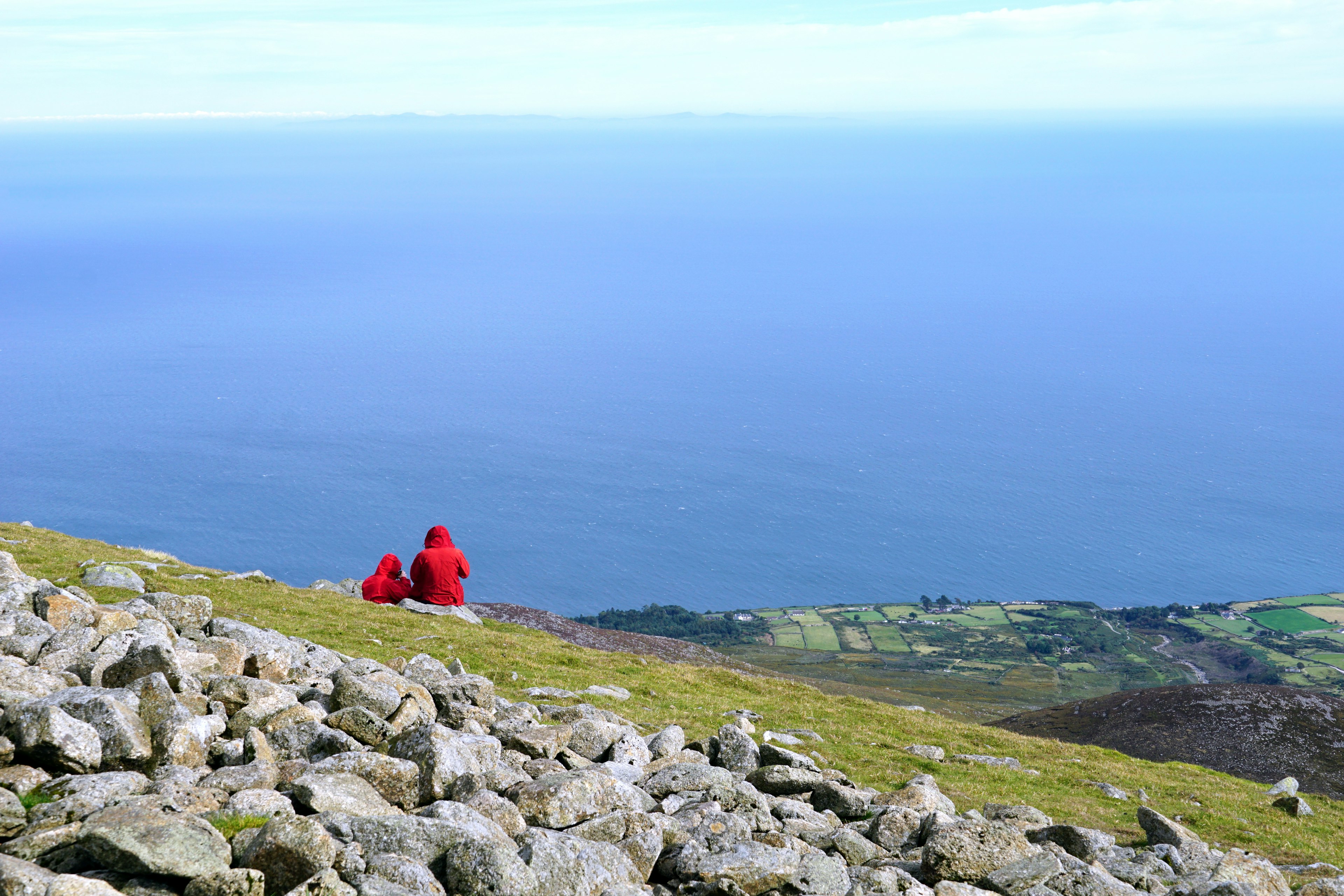 Two people wearing red coats with their hoods pulled over their heads sit on a rock on a moutaintop looking out to sea