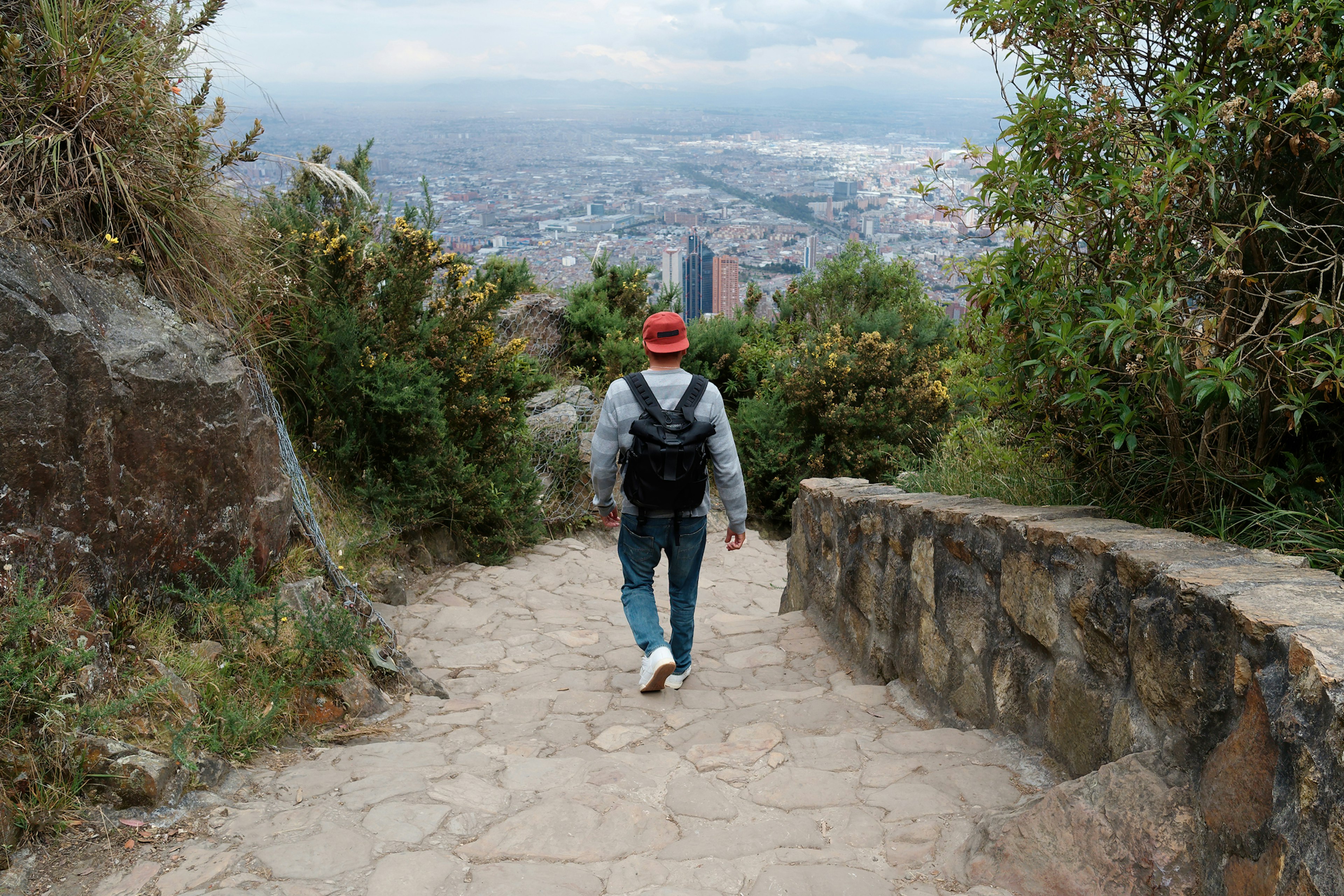 A man seen from behind walking a steep stone path down a mountain to the city below