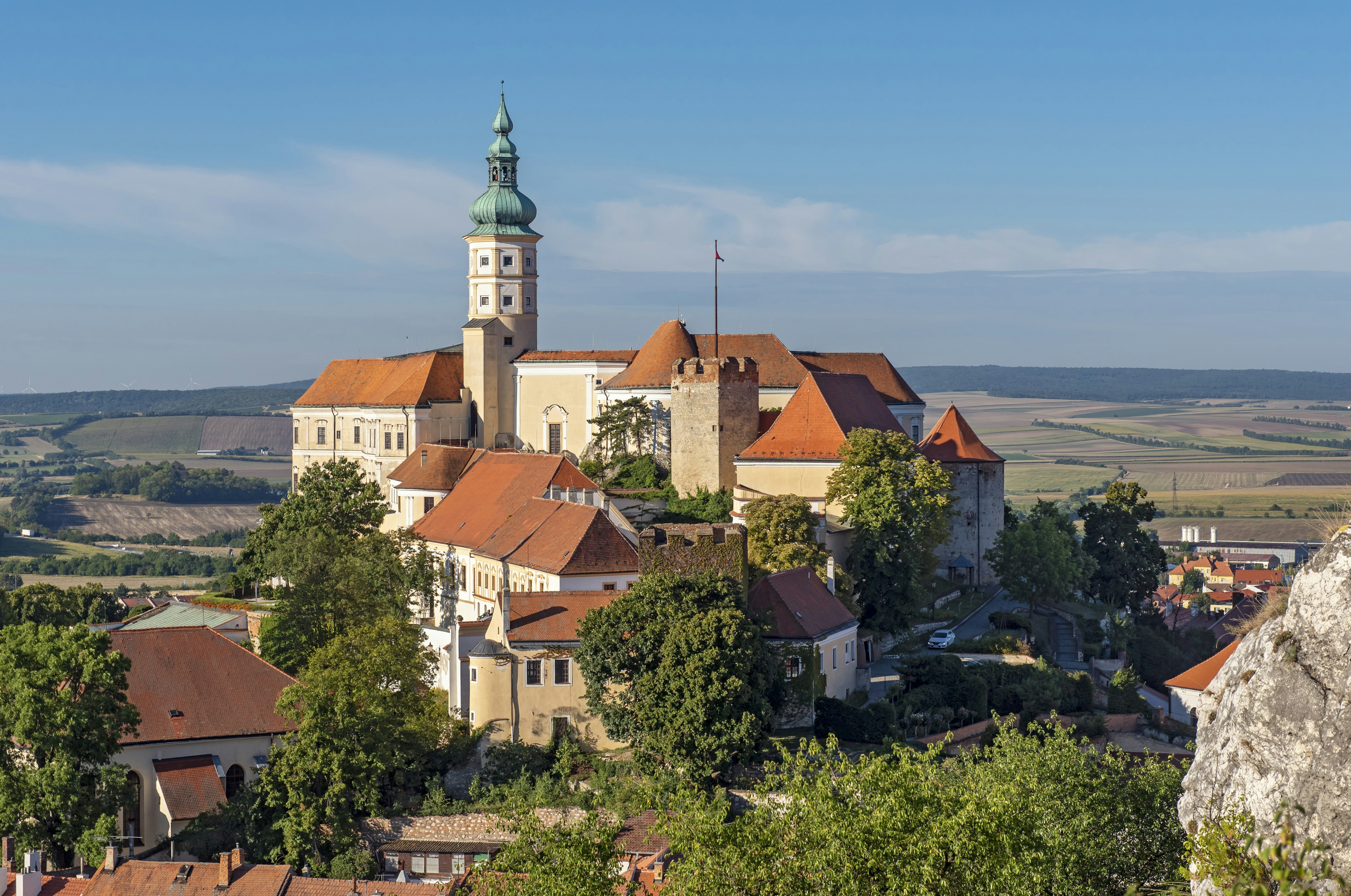 A red-roofed chateau with a tall steeple rises on cliffs over a village and vineyards in the countryside