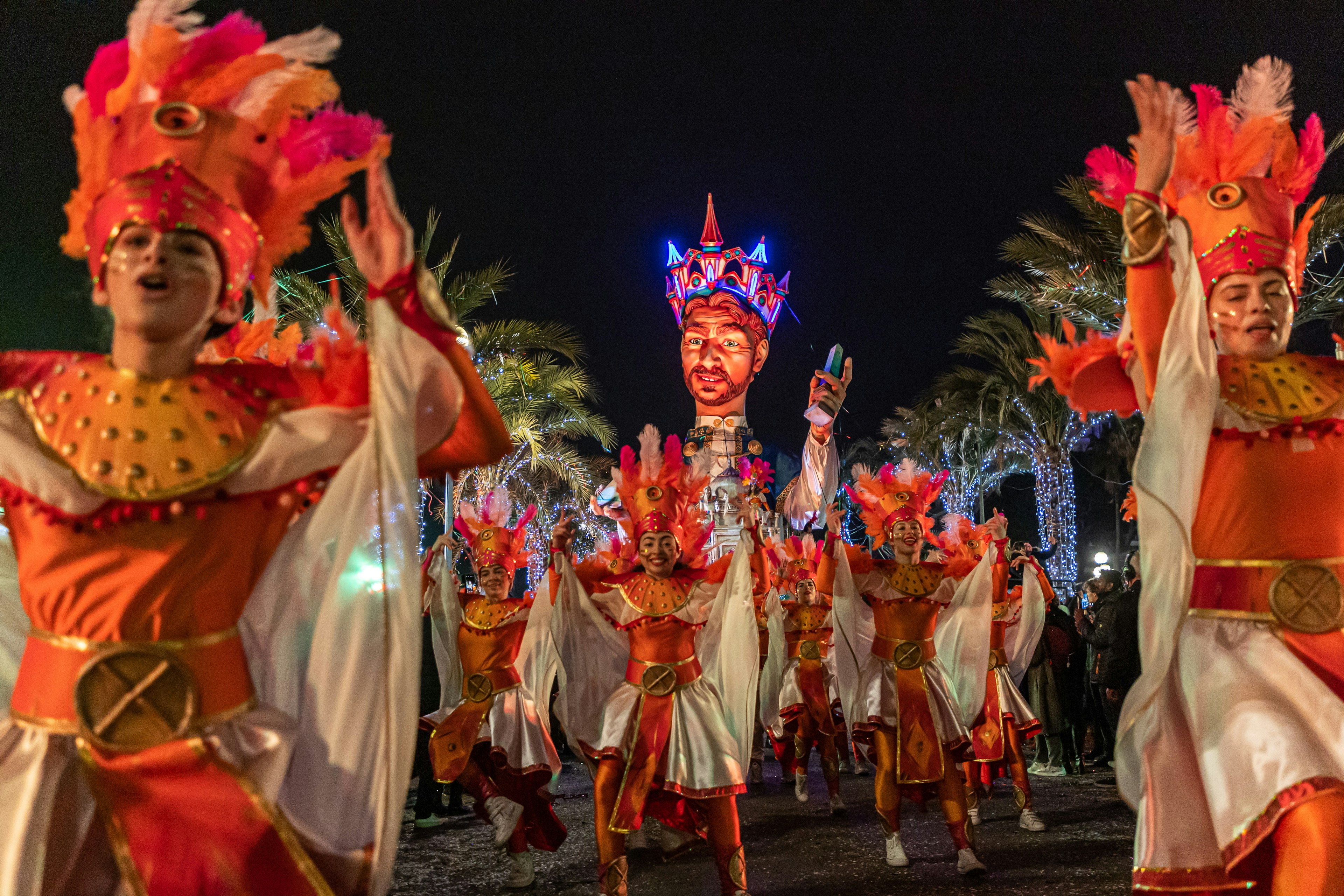 People in bright-orange costumes dance in formation against a row of illuminated palm trees