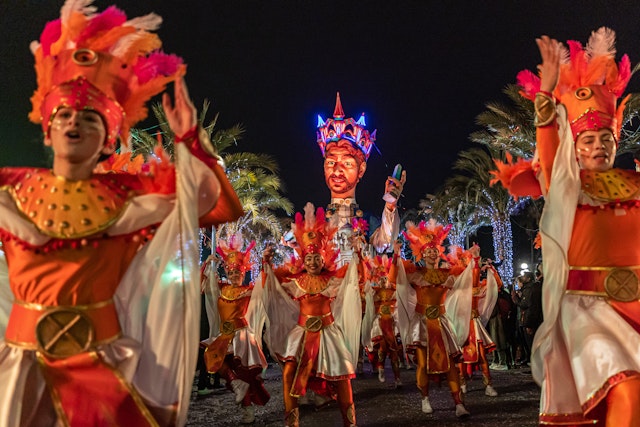 People in bright-orange costumes dance in formation against a row of illuminated palm trees