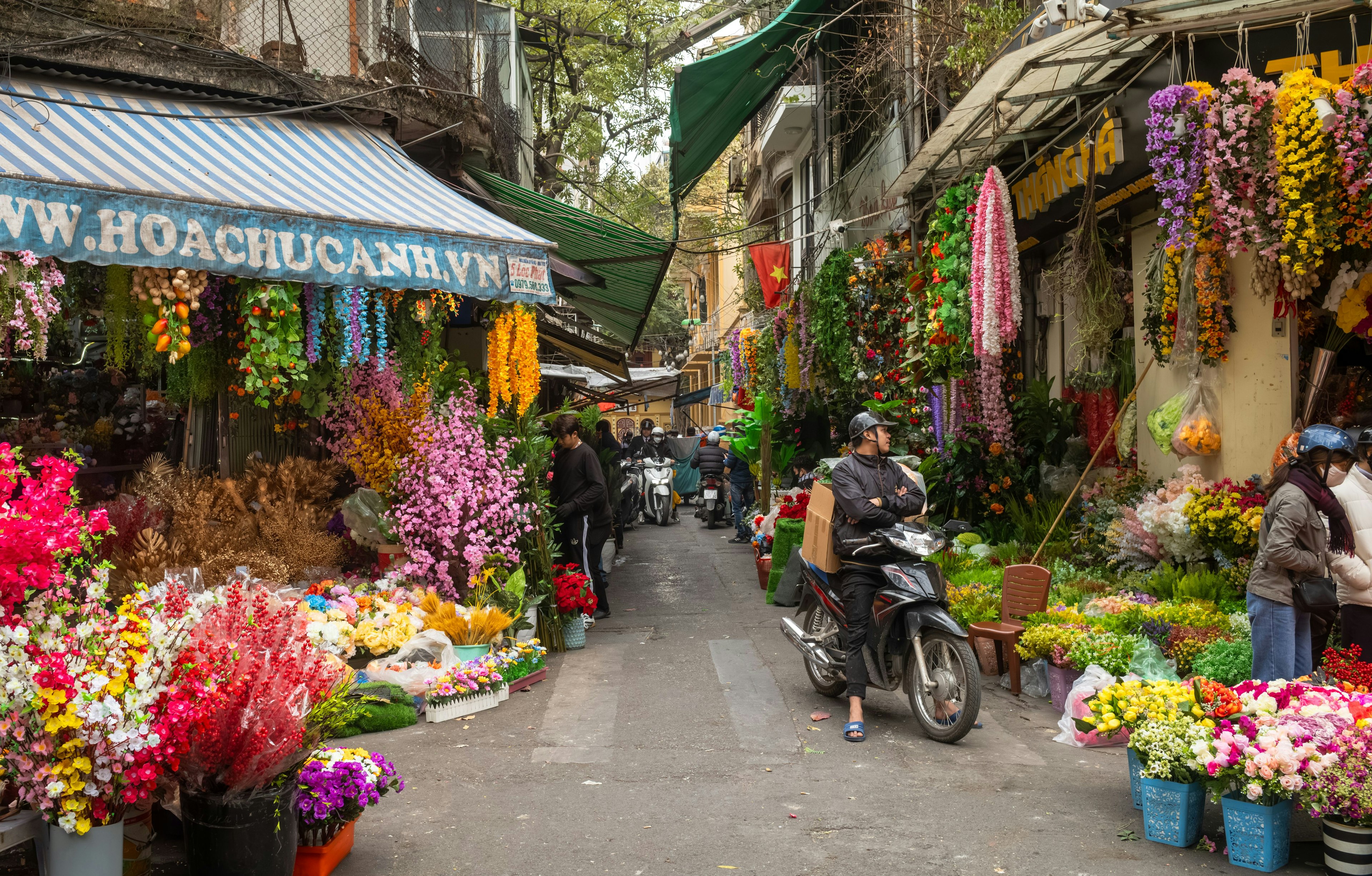 A man on a motorbike waits by shops on either side of an alley selling artificial multi-colored flowers in the Old Quarter of Hanoi, Vietnam